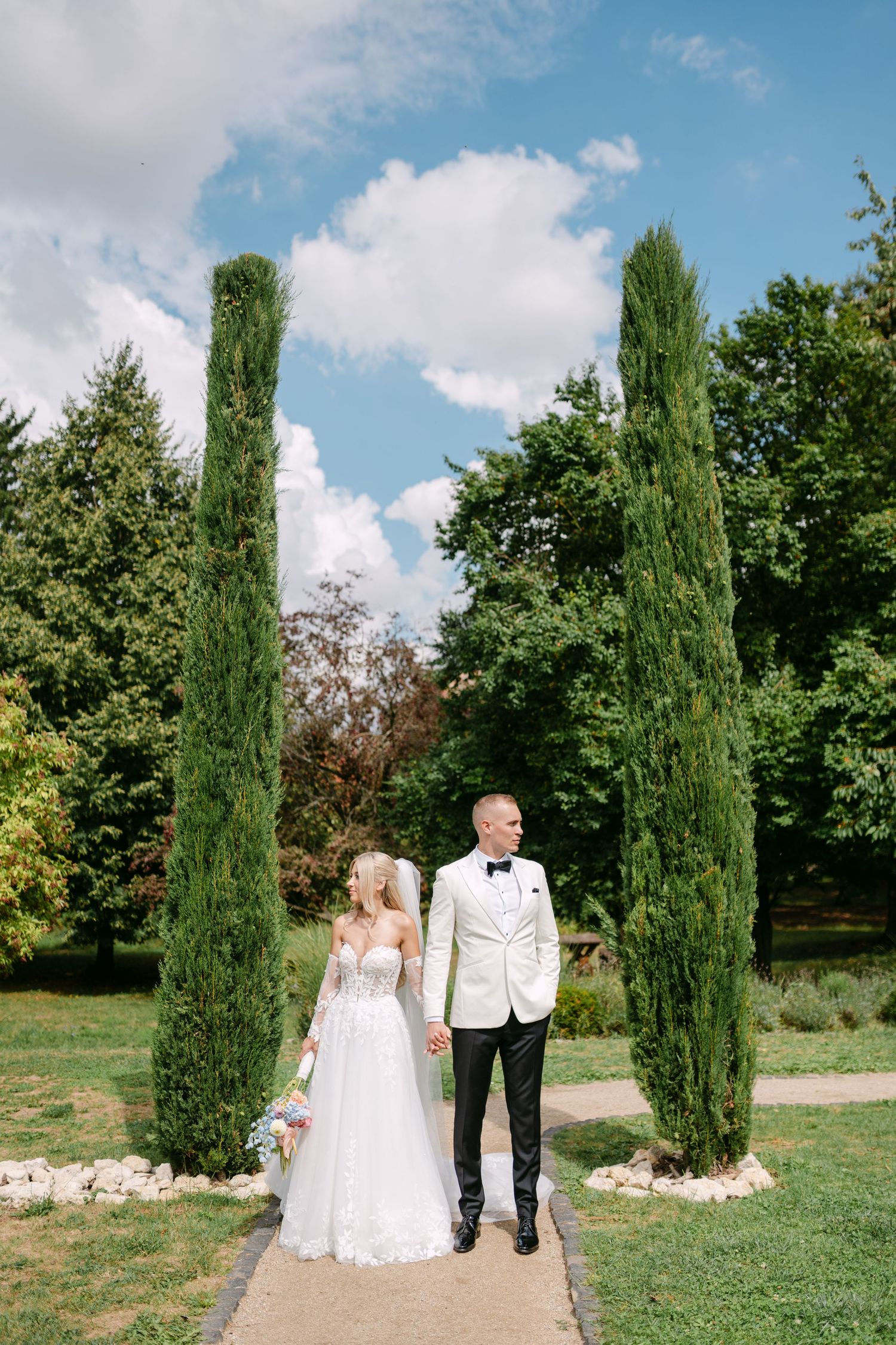 Elegant wedding couple walks along a garden path lined with tall cypress trees.