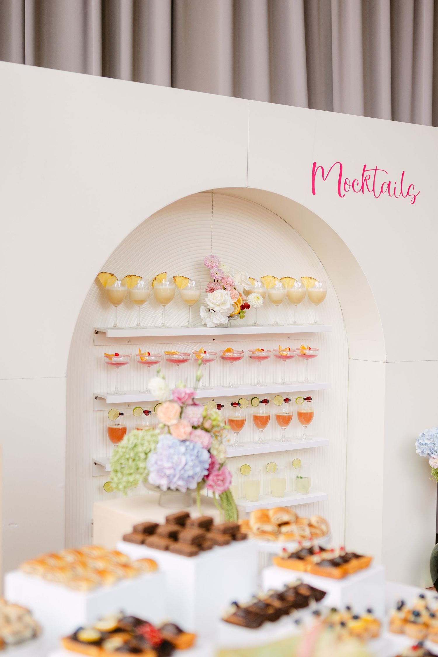 Wedding dessert display featuring mocktails and sweets arranged on elegant white shelving.