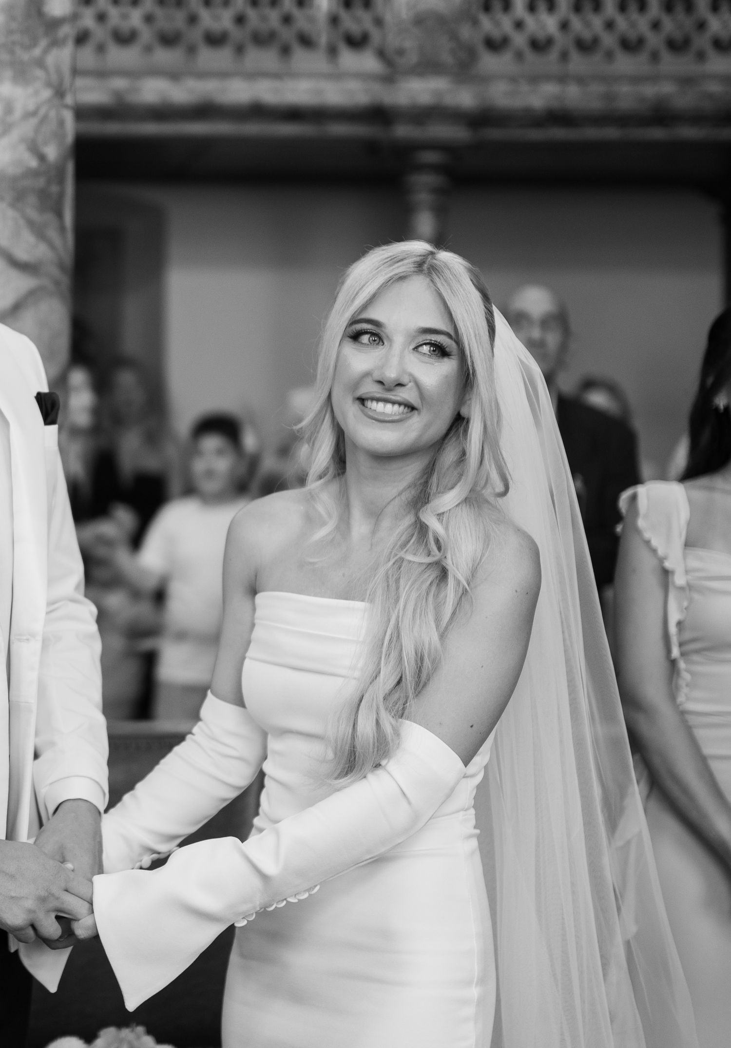 Bride in a long-sleeved white wedding dress smiles during an emotional moment at her wedding celebration.