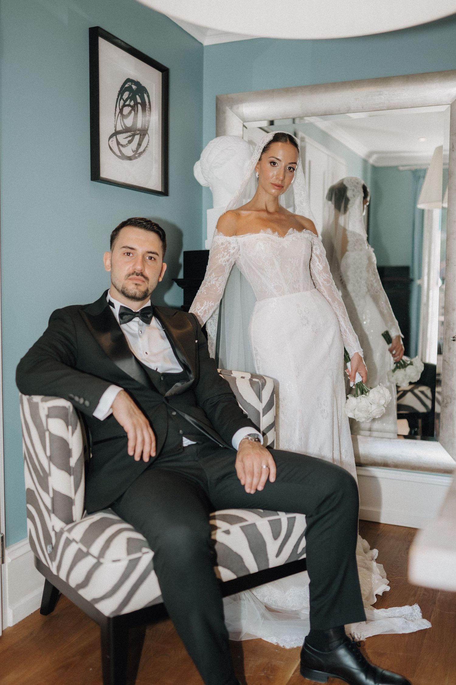 Wedding photoshoot featuring a classic black tuxedo and white bridal gown in a blue-walled room.