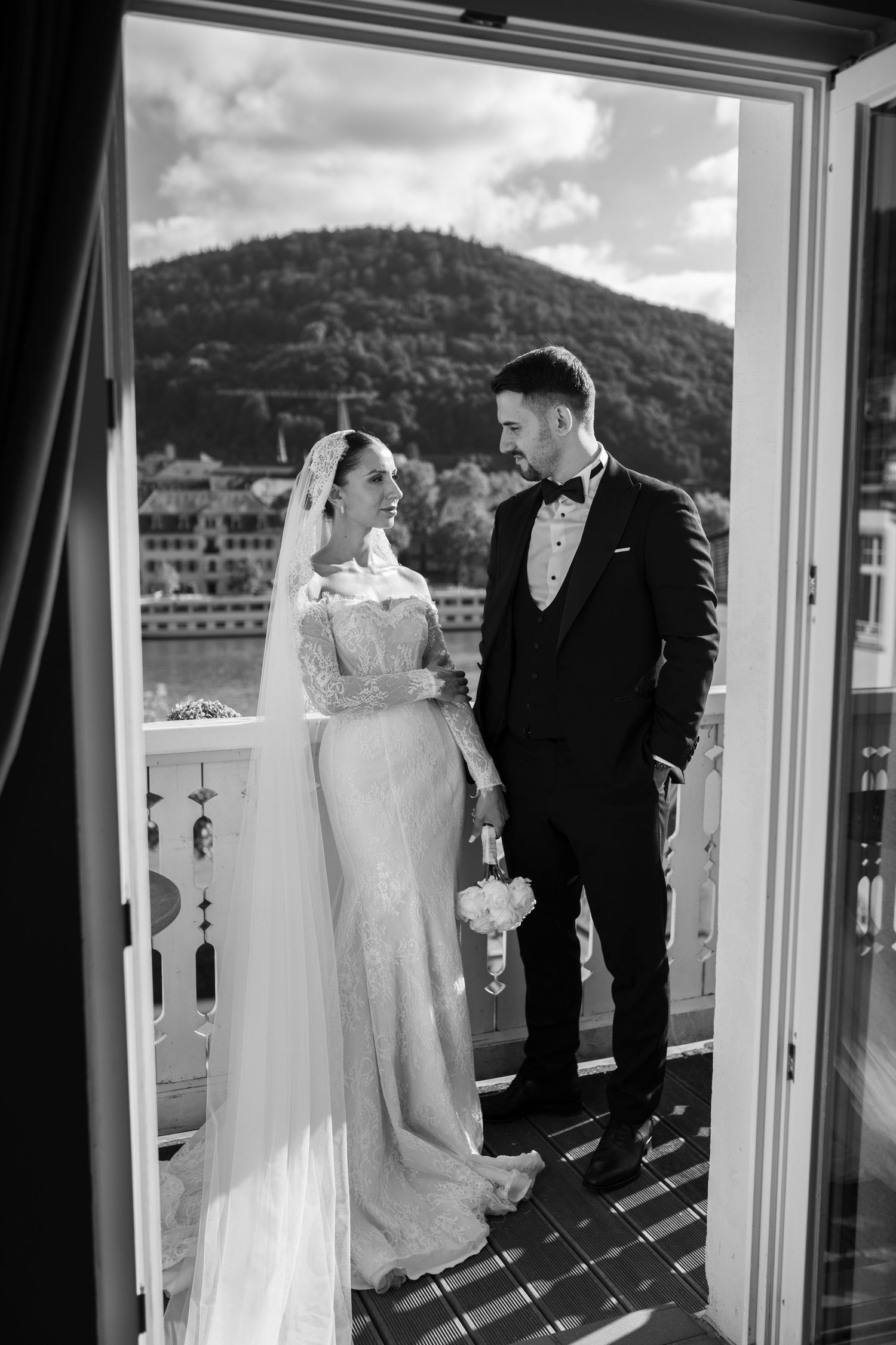 Wedding couple in formal attire standing by large windows with mountain views.
