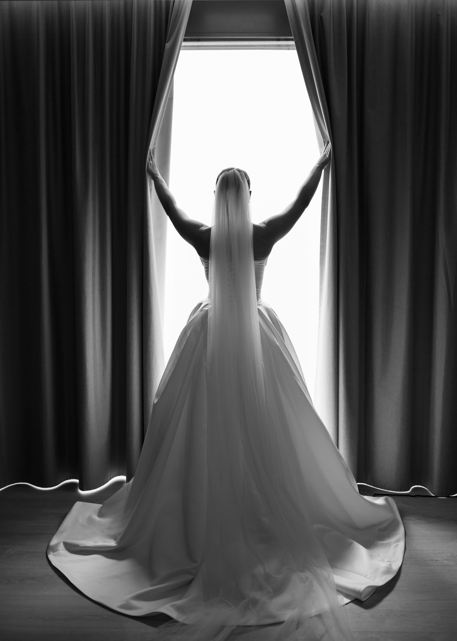 Dramatic silhouettes of wedding dress against window light and dark curtains.