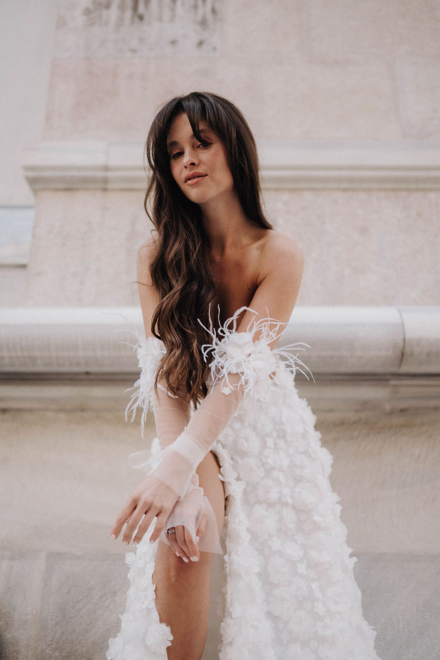 Fashion model posing in delicate white feathered dress against stone architecture.