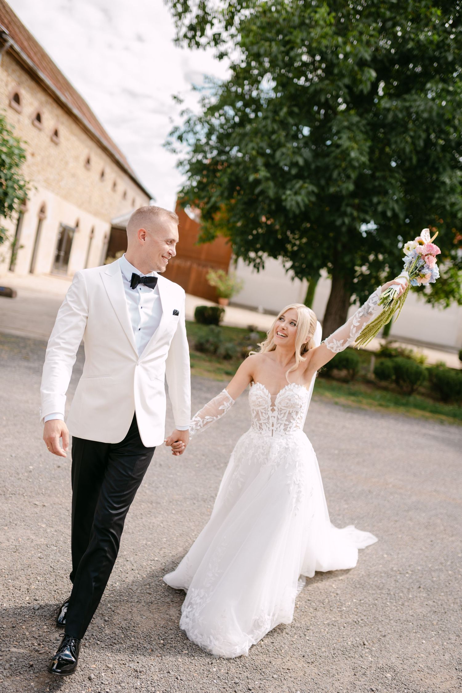 Playful wedding couple dancing and twirling on a gravel path outside historic buildings.