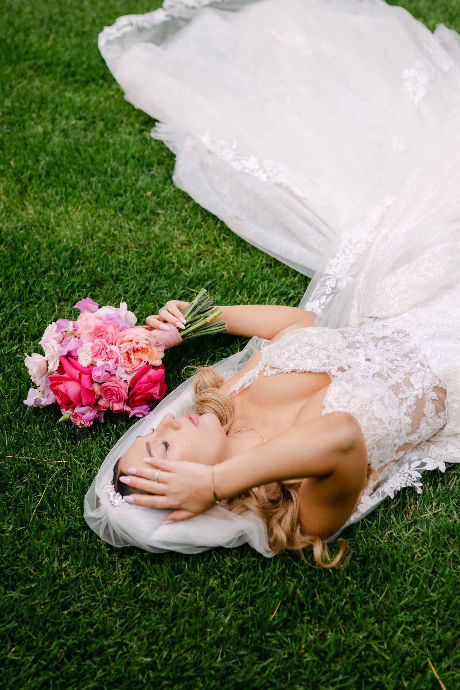 A bride in lace dress lying on grass with pink bouquet in artistic wedding photo.