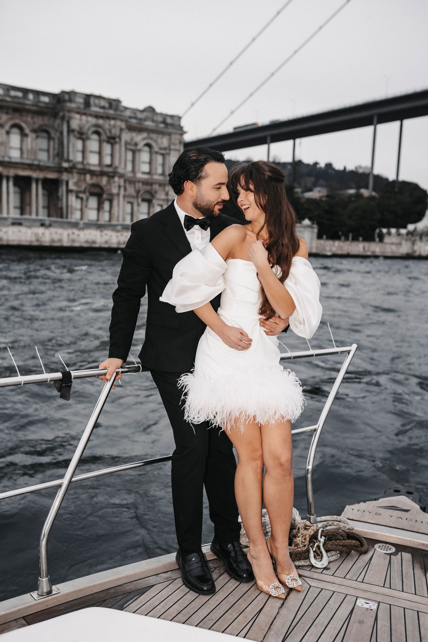 Intimate moment between couple on yacht deck with waterfront architecture in background.