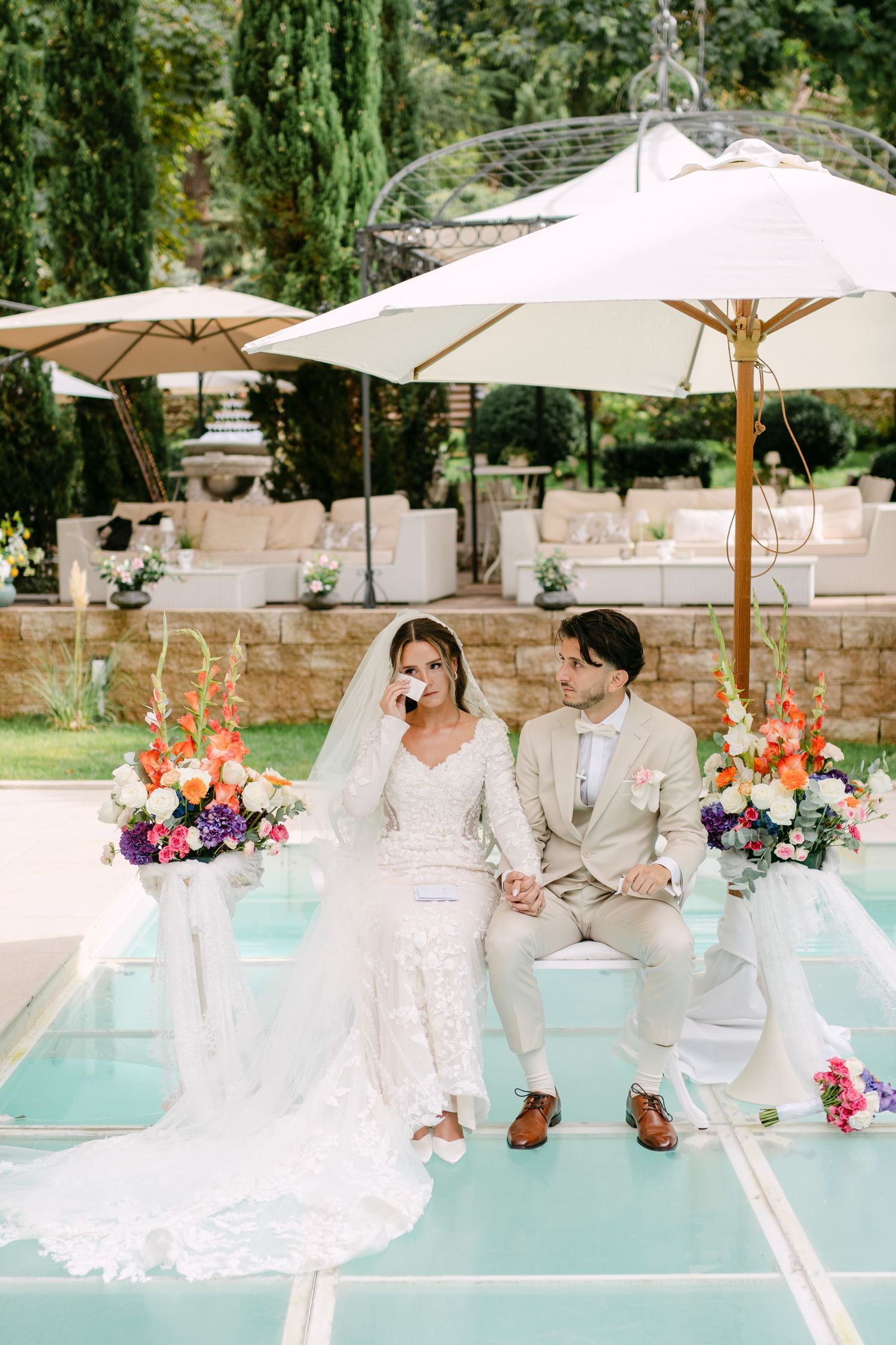 Elegant poolside wedding ceremony setup with white umbrellas and colorful floral arrangements.