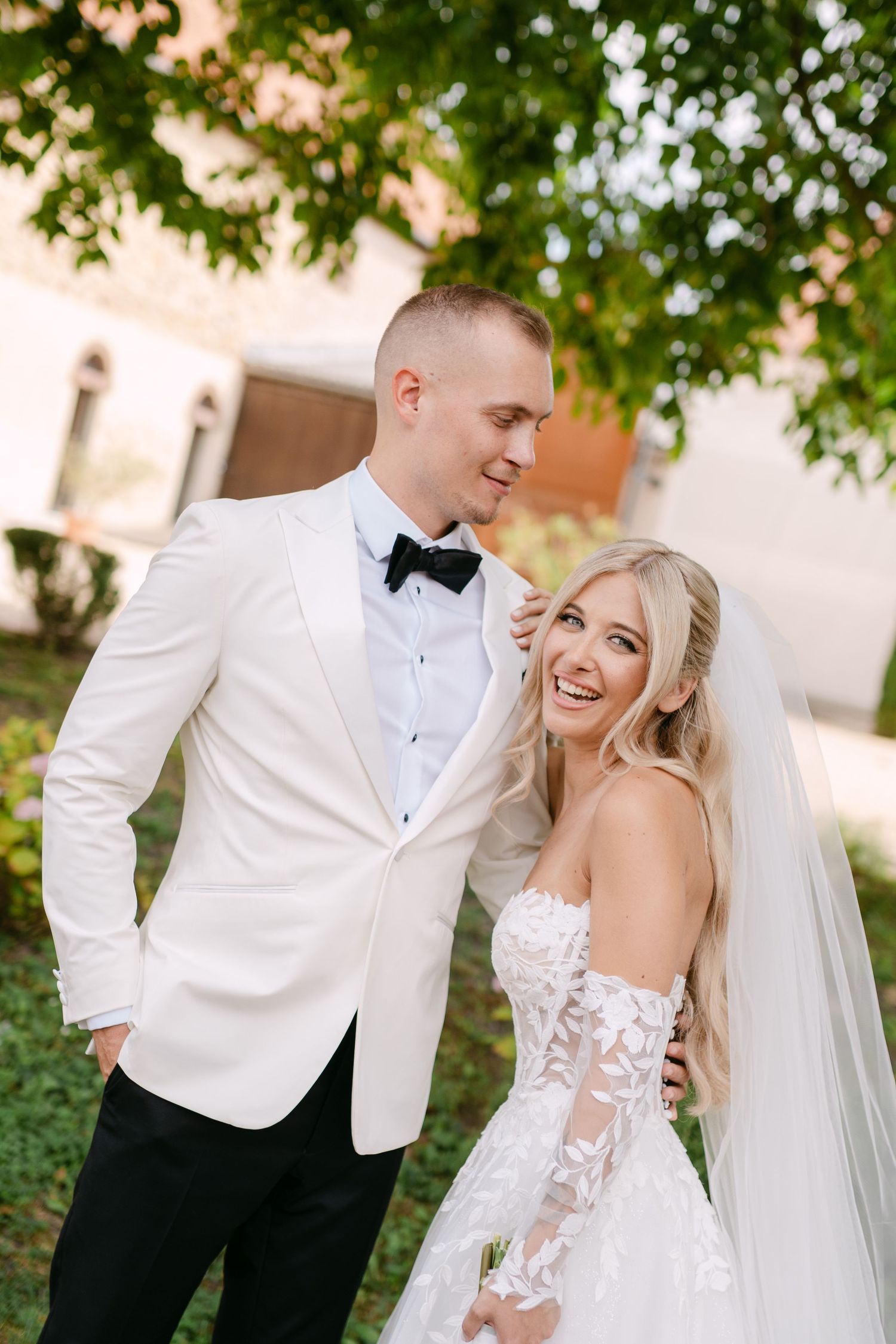 A joyful wedding couple shares intimate moments under green trees in a garden setting.