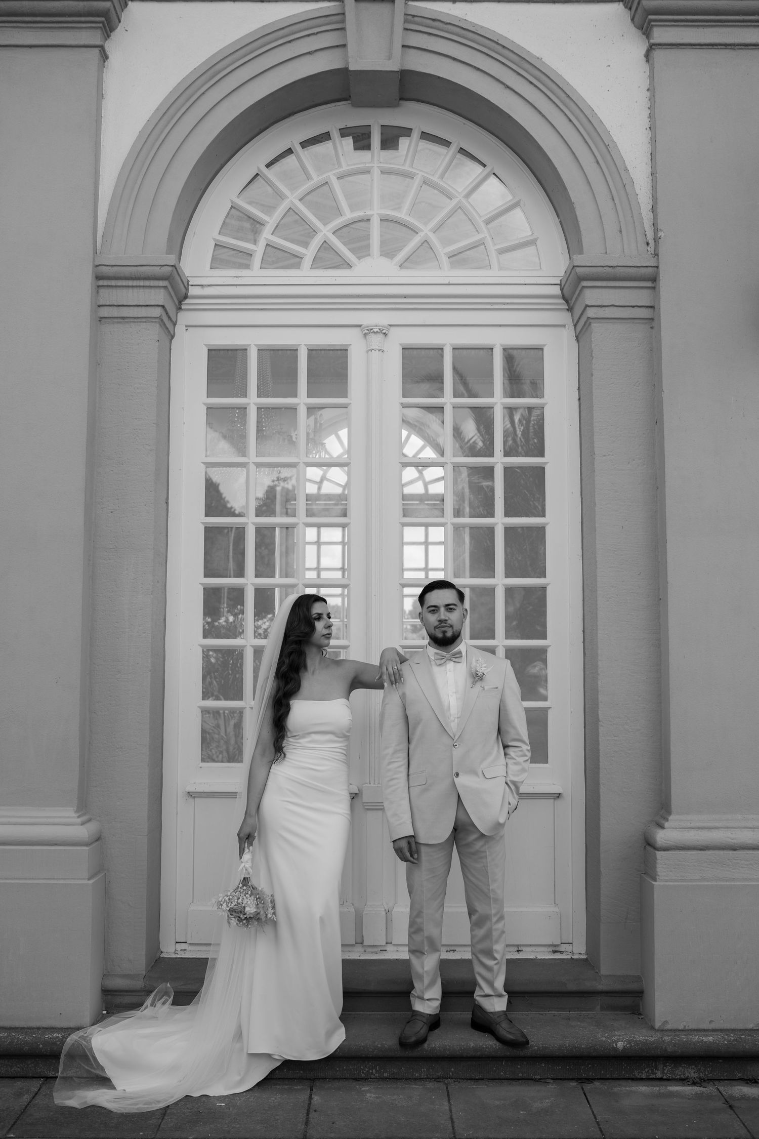 A wedding couple poses elegantly against arched windows in a classic architectural setting.