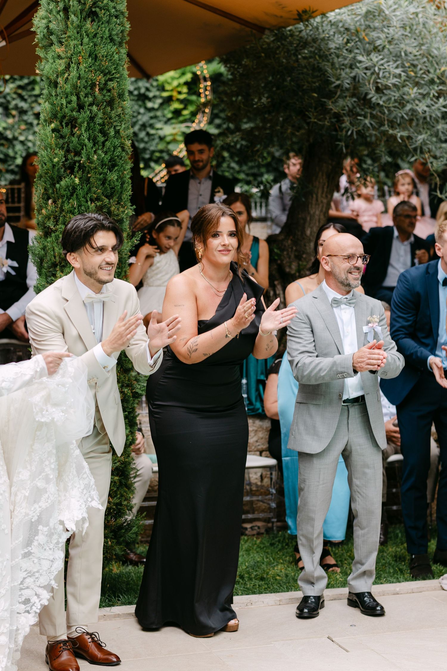 Wedding guests applauding during outdoor ceremony by cypress trees.