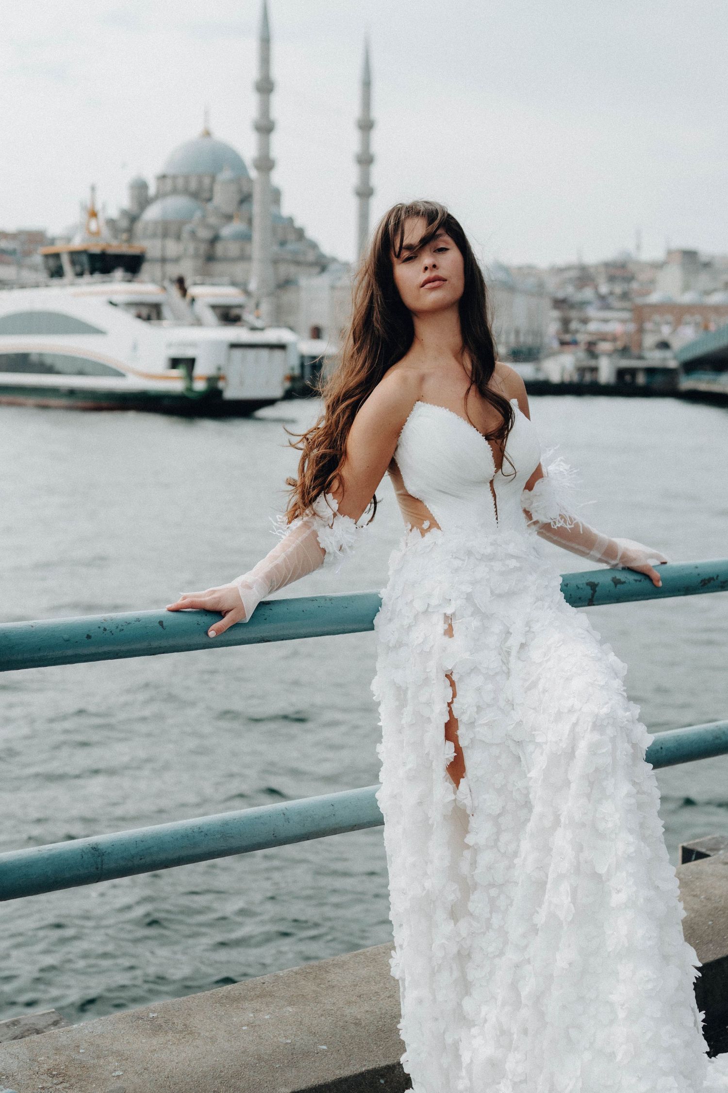Bride in white dress poses by waterfront with Istanbul mosque skyline in background.