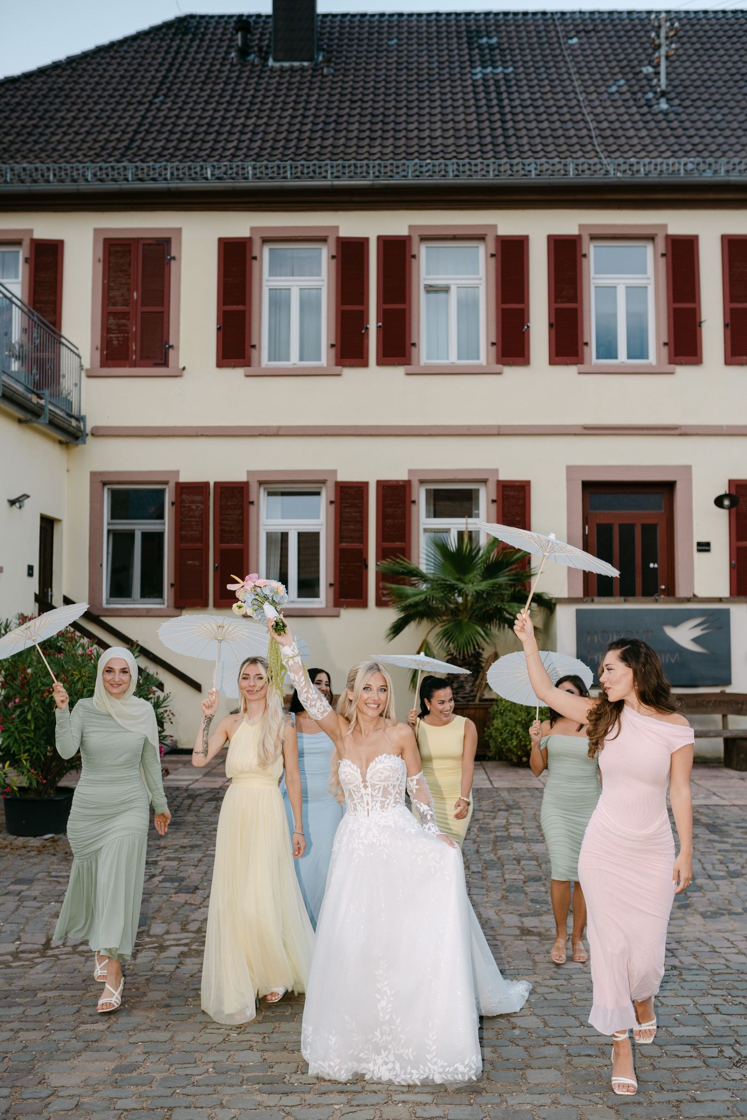 Wedding party in pastel dresses poses together outside traditional European-style building.