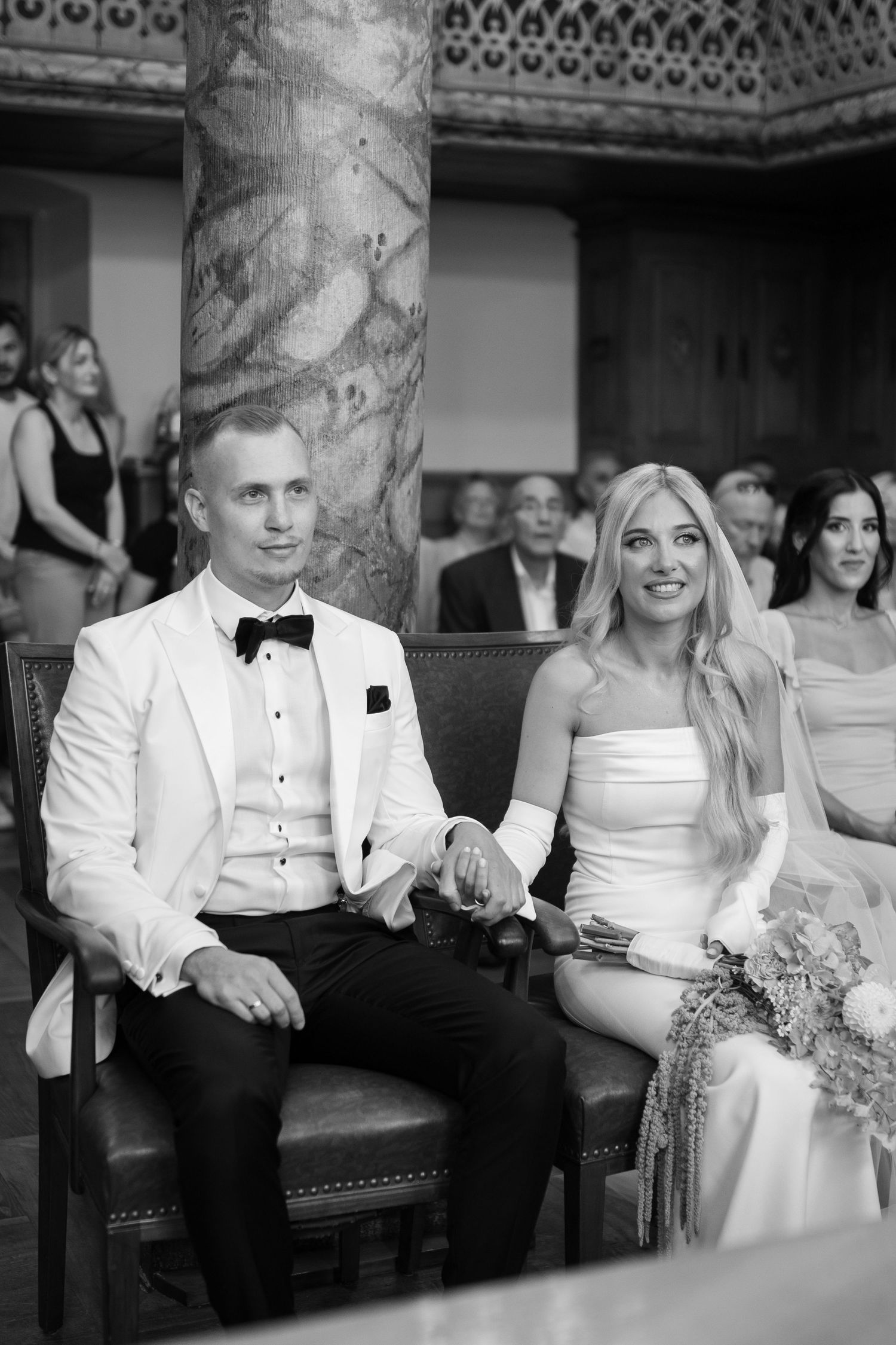 Wedding guests seated in formal attire at an elegant venue with ornate architectural details.
