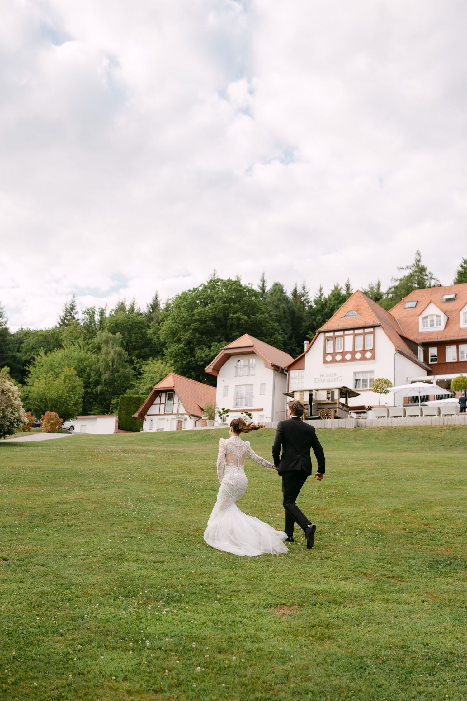 Couple runs through a scenic field with houses and trees in the background during their wedding.