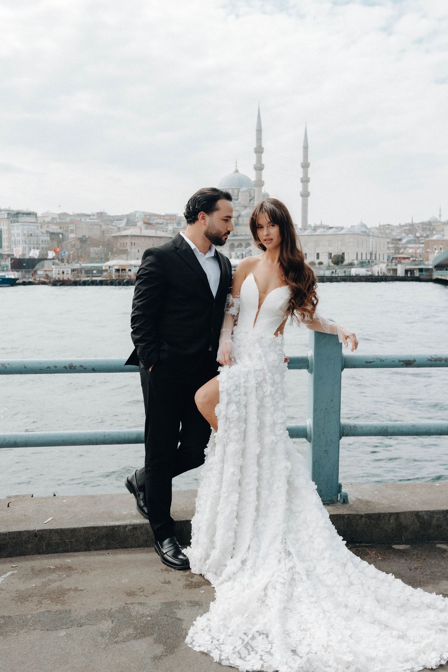 Wedding couple poses along waterfront with Istanbul mosque and cityscape in background.