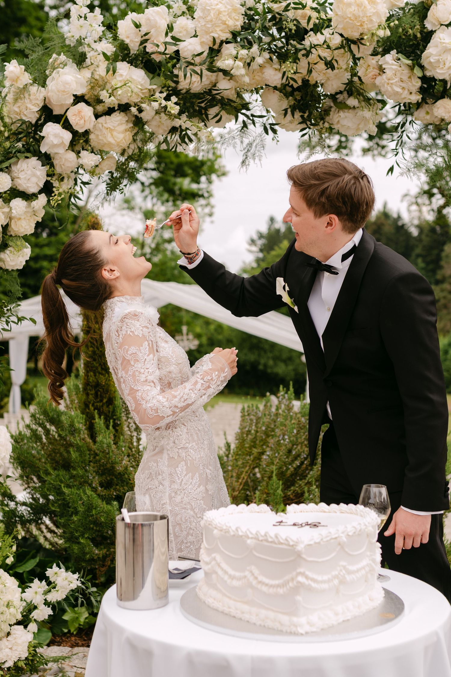 Couple shares a joyful moment cutting their wedding cake under a floral arch decorated with white roses.