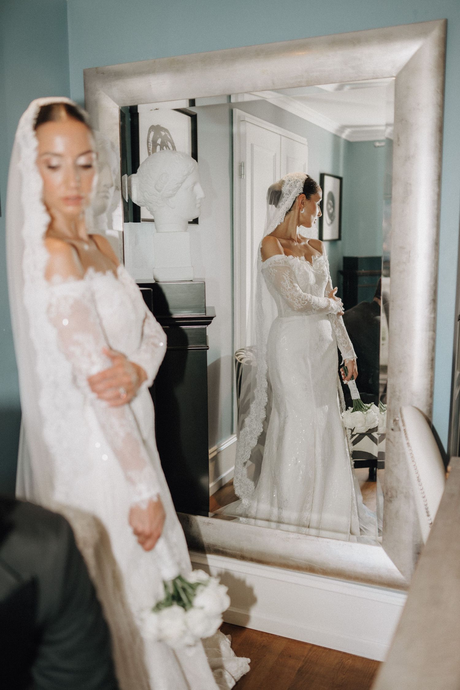 Elegant wedding dress reflection in a large mirror showing intricate lace details and flowing white fabric.