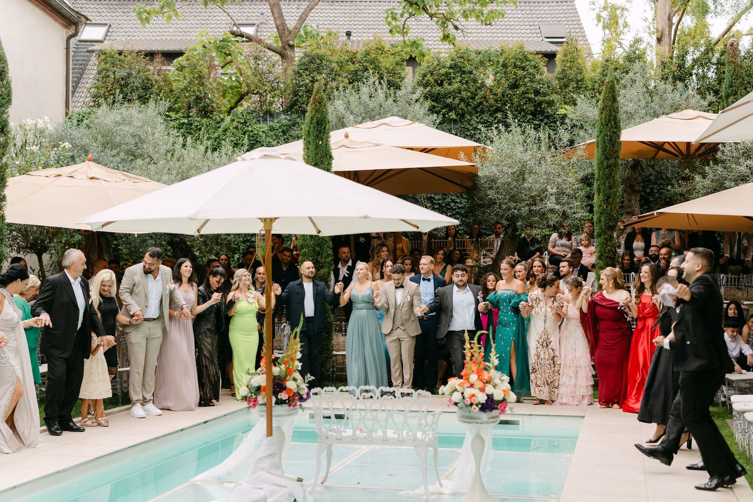 Wedding guests gathered around pool for outdoor ceremony with white umbrellas and floral decor.