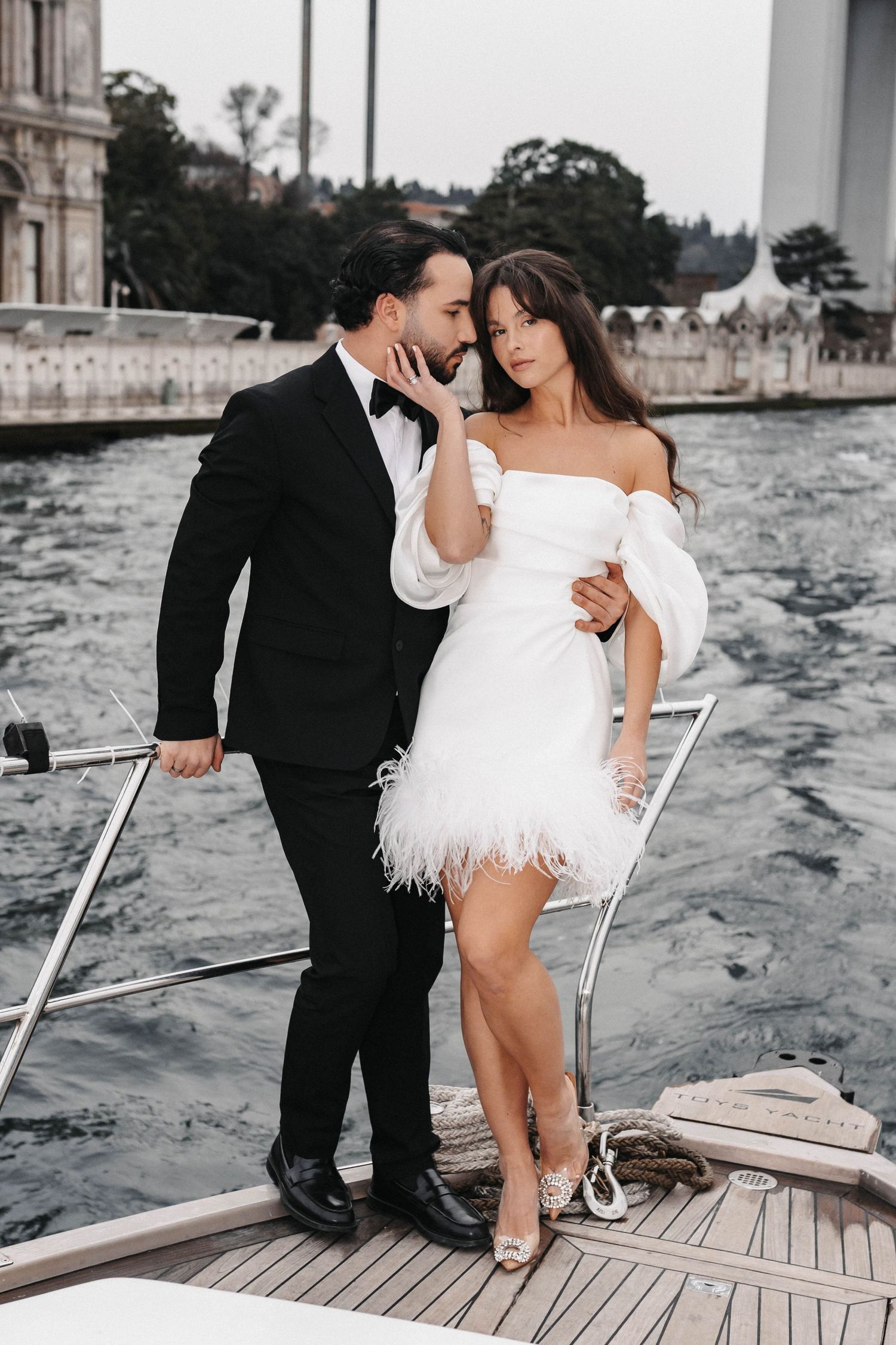 Elegant couple embraces on yacht deck with waterfront backdrop.