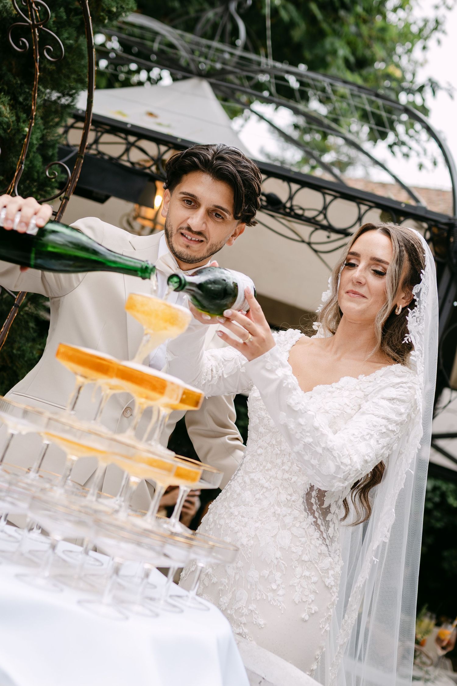 Couple pouring champagne into a tower of crystal glasses at an outdoor wedding reception.