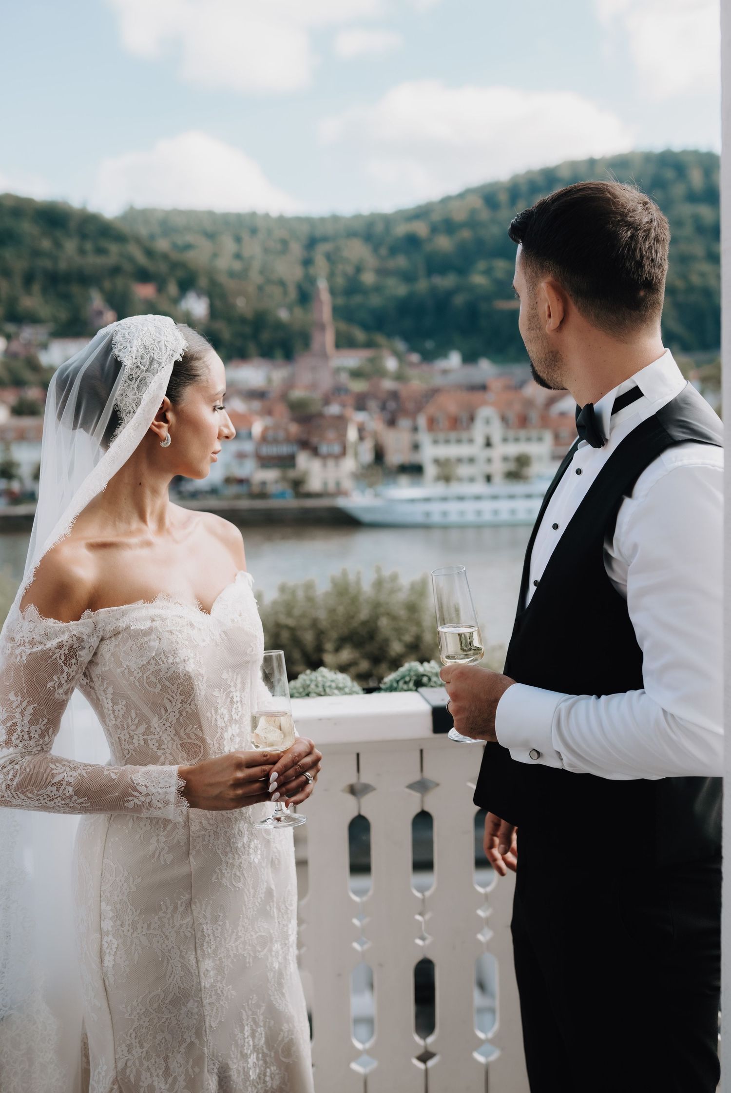 Elegant couple sharing drinks on a balcony with mountain and river views in the background.
