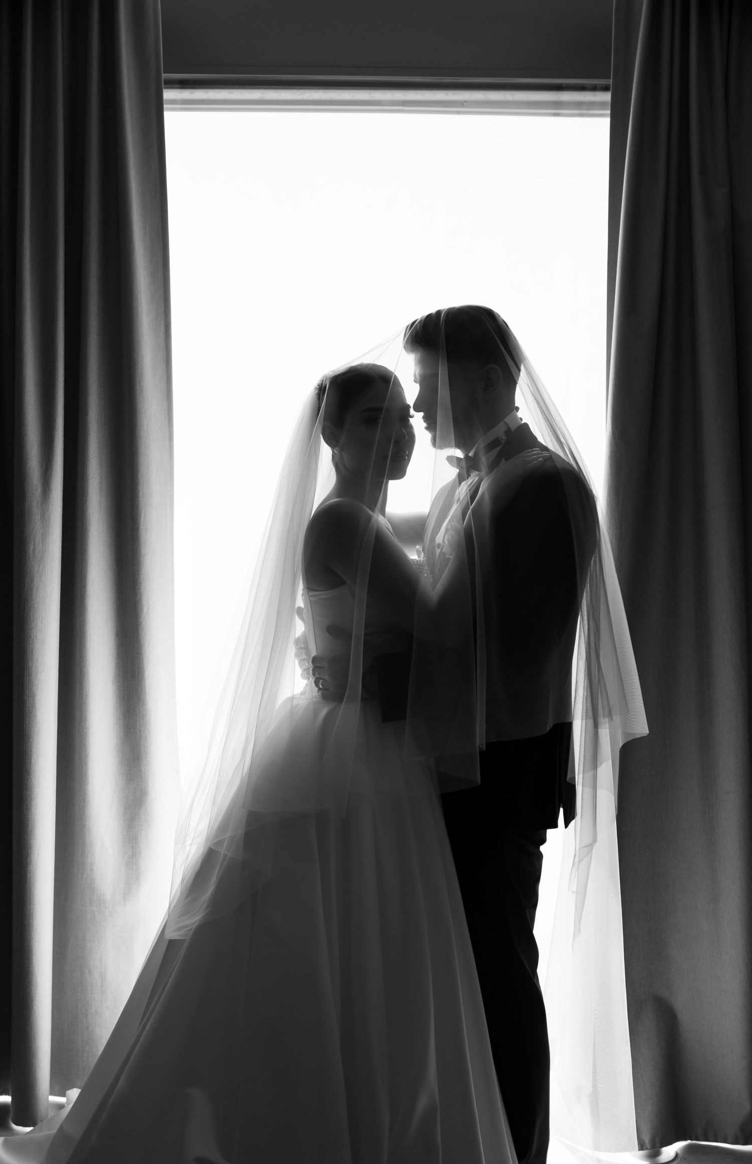 Silhouetted figures of bride and groom against window with flowing curtains.