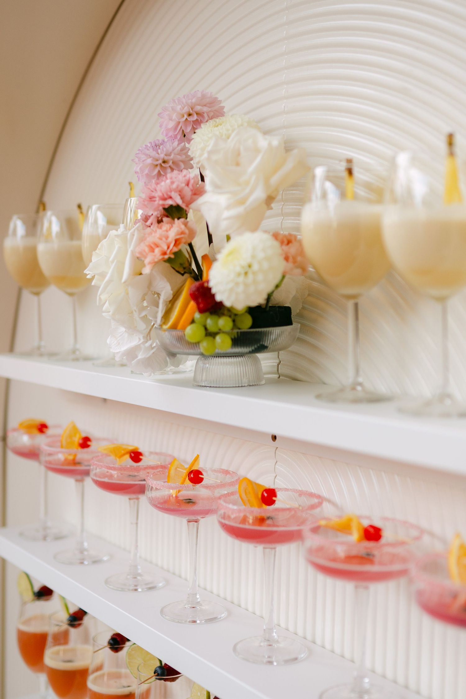 Row of colorful cocktails garnished with flowers displayed on white shelving at a wedding bar.