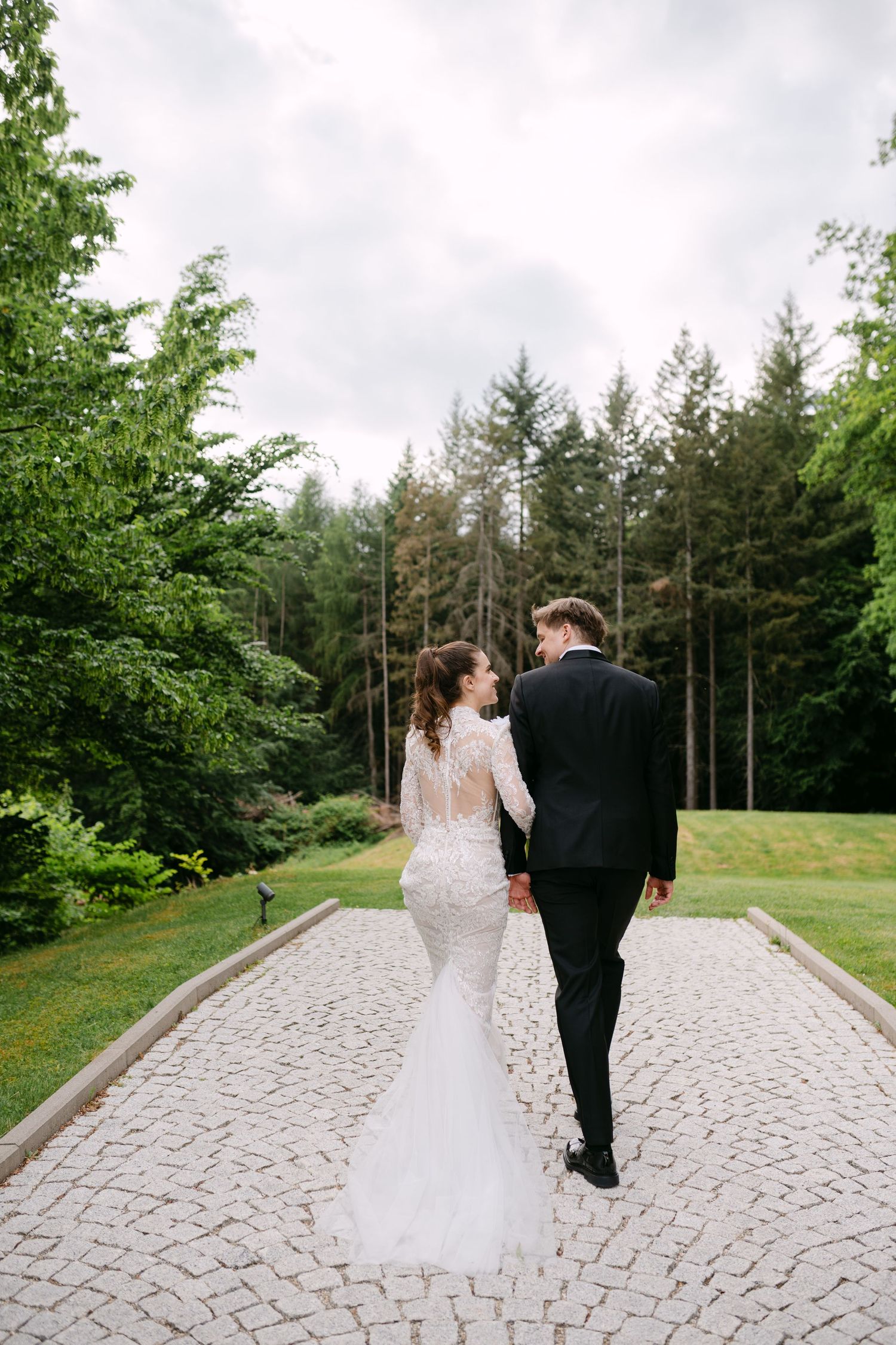 Newlyweds walk along a stone pathway surrounded by lush greenery and pine trees.