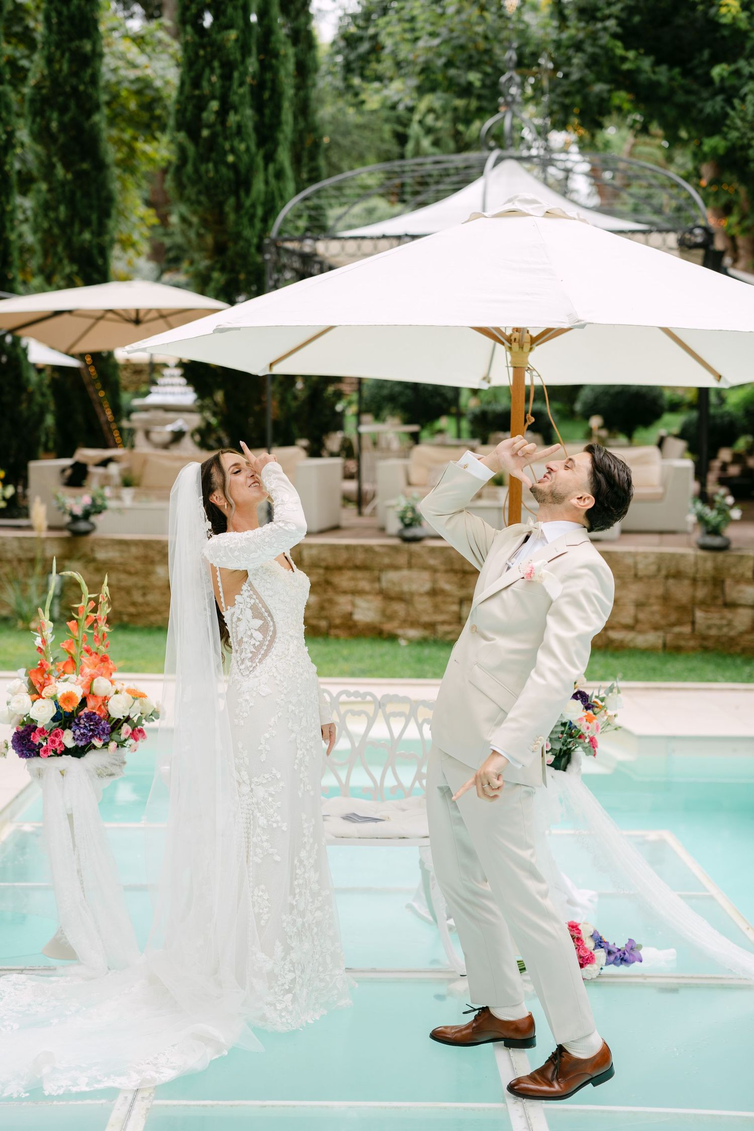 Playful wedding couple posing under white umbrellas by pool with floral decorations.