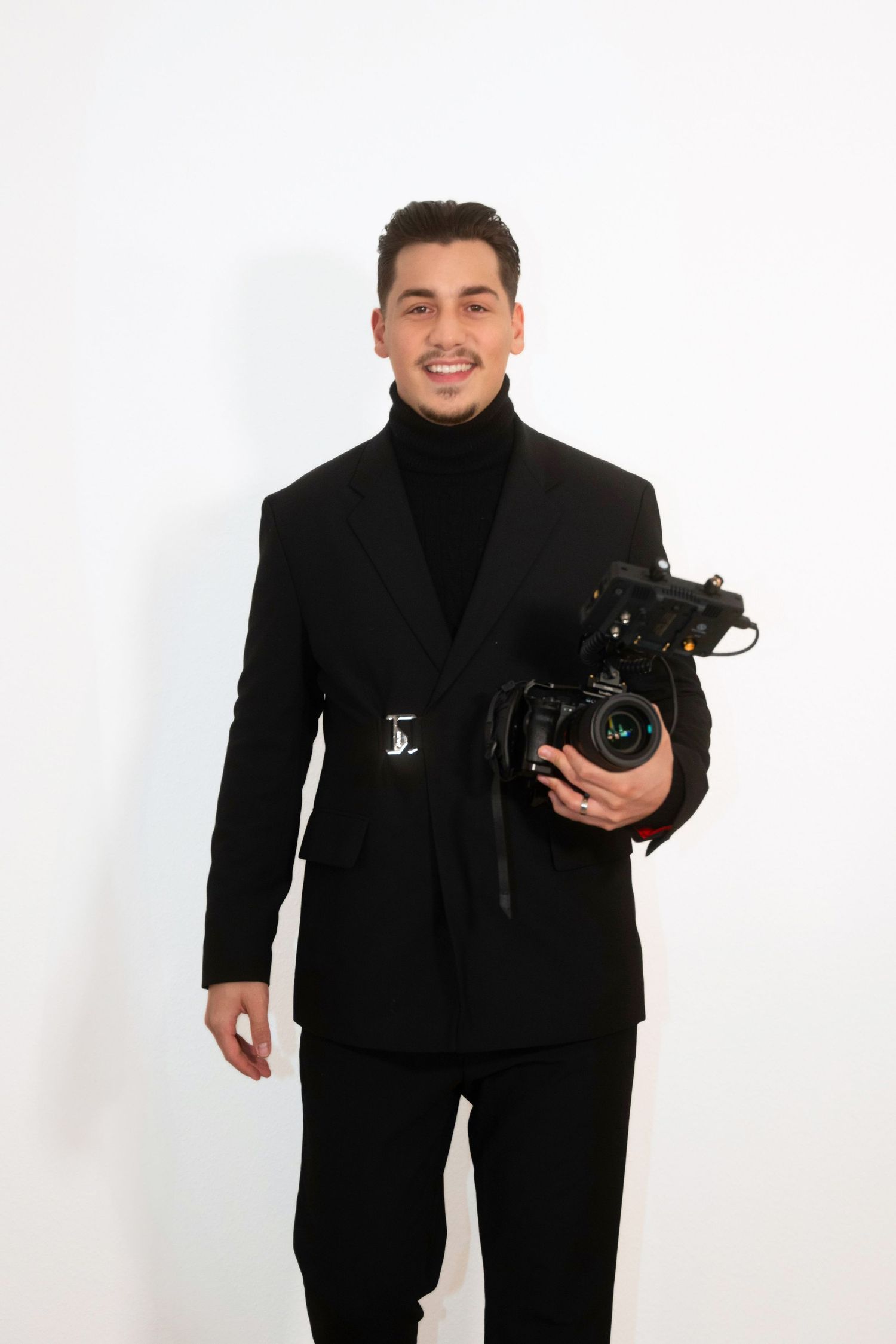 Professional photographer in all black attire holding camera equipment against white background.