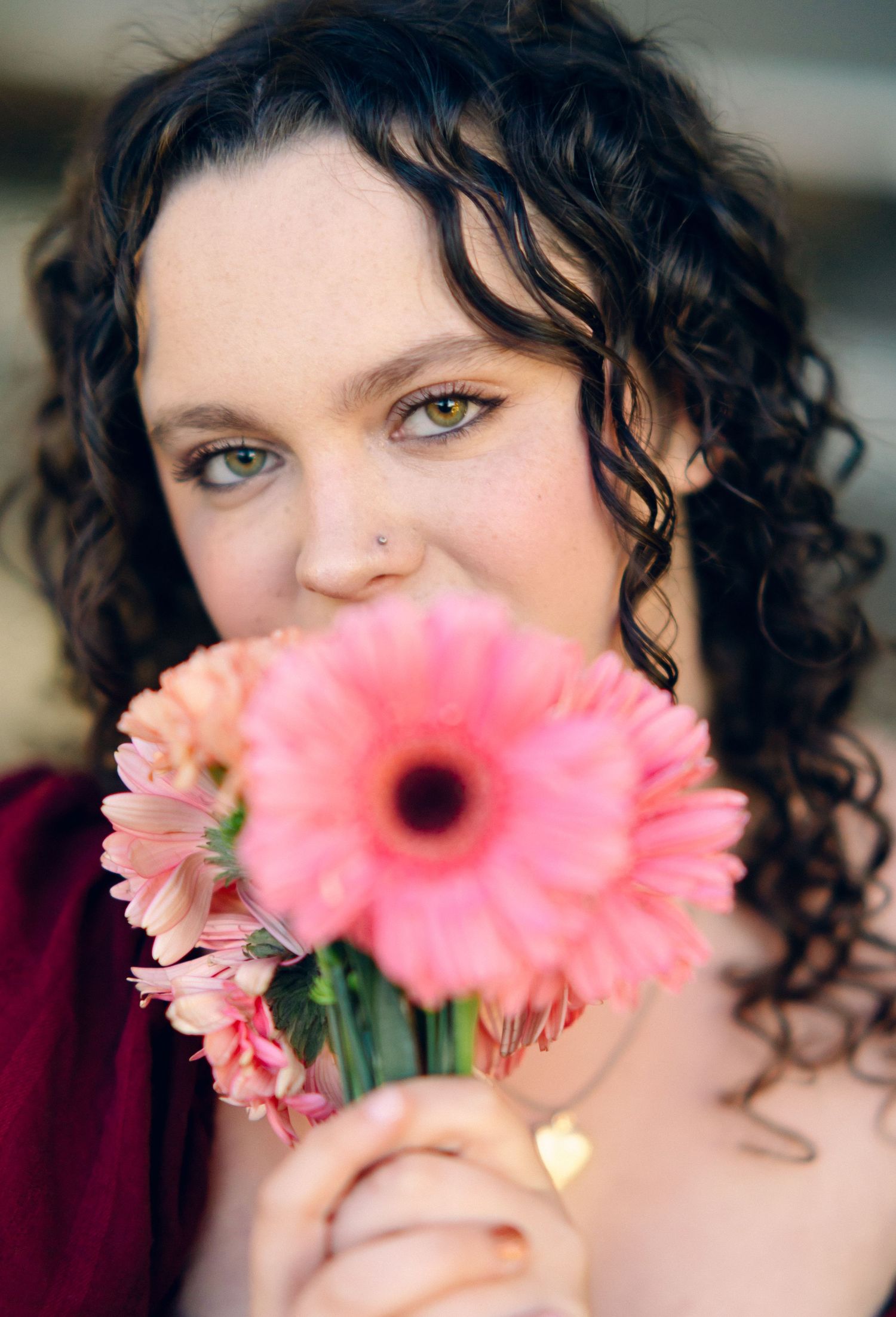 Woman with curly hair holds pink gerbera daisies near her face, wearing a burgundy dress with gold ring.