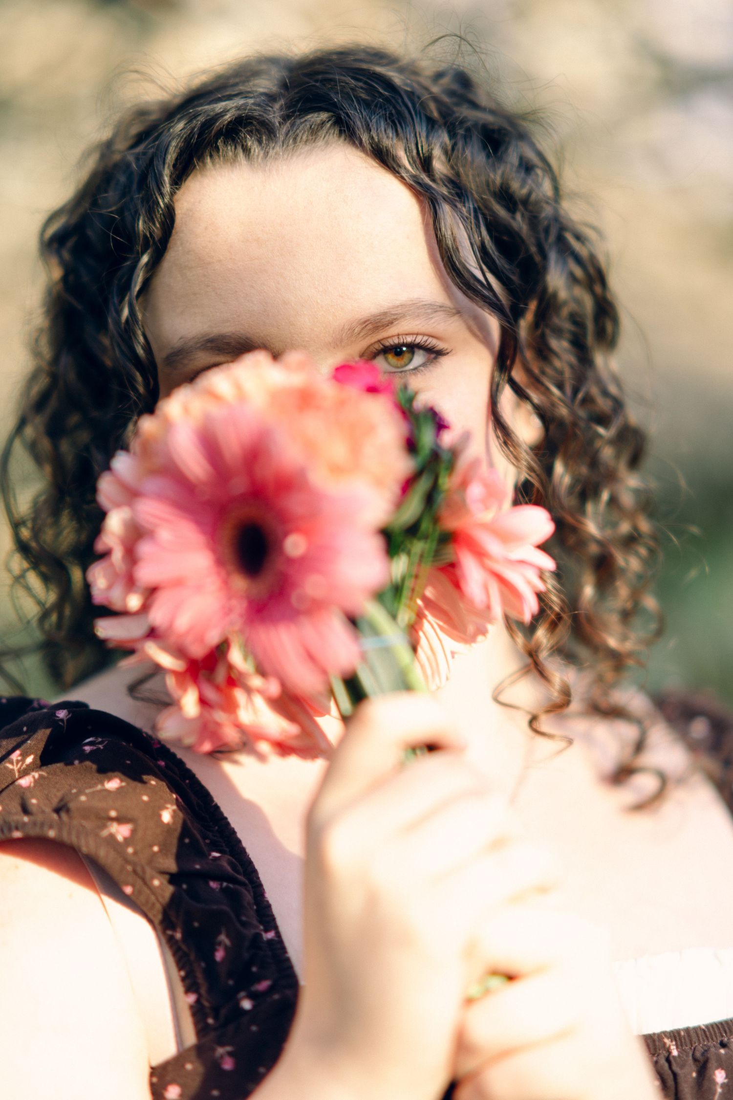 Young woman with curly dark hair holding pink gerbera daisies in front of her face outdoors.