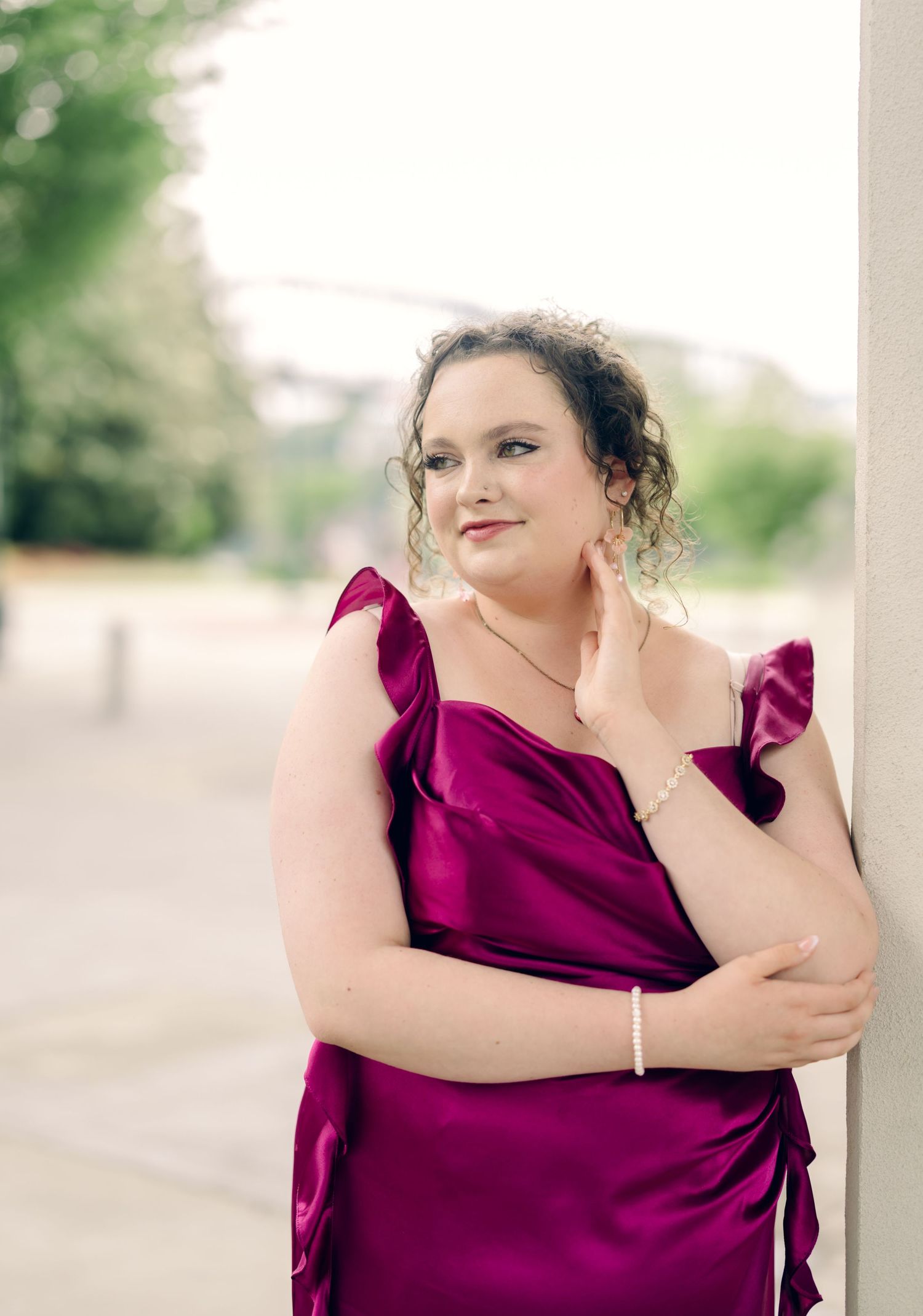 Young woman in elegant magenta satin gown posing gracefully against a pillar outdoors.