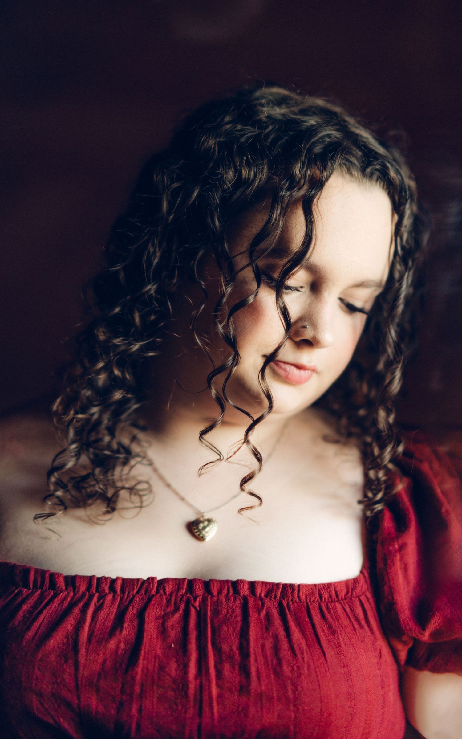 High school senior with curly dark hair wearing a red off-shoulder top and gold heart necklace, looking down softly.