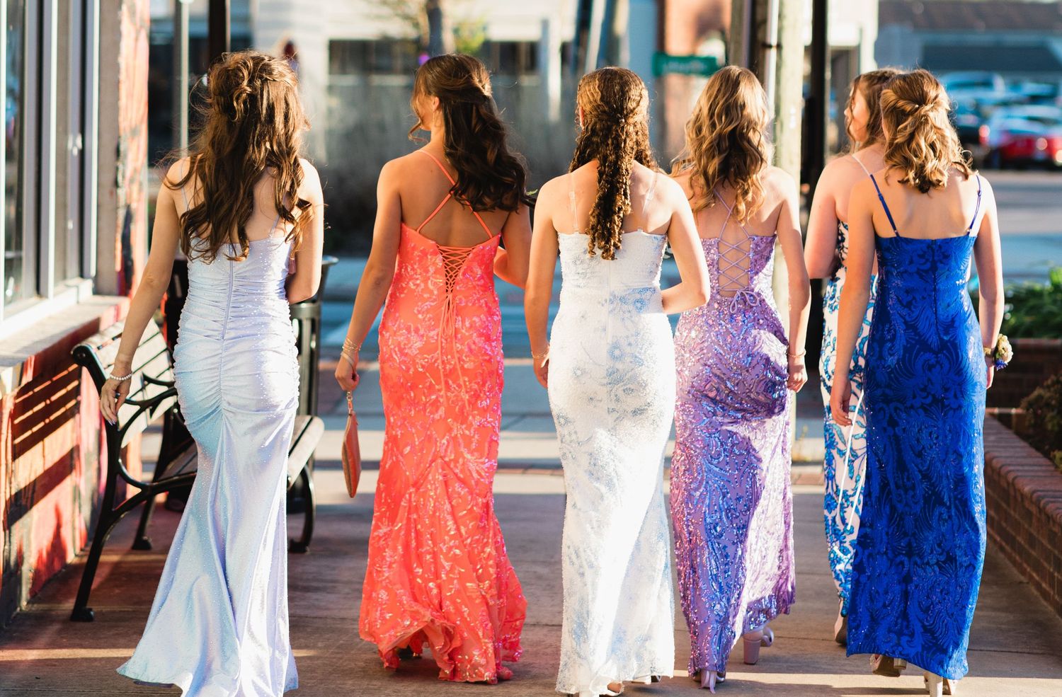 Five young women in elegant formal gowns walk together on a sidewalk, viewed from behind, wearing colorful prom dresses.