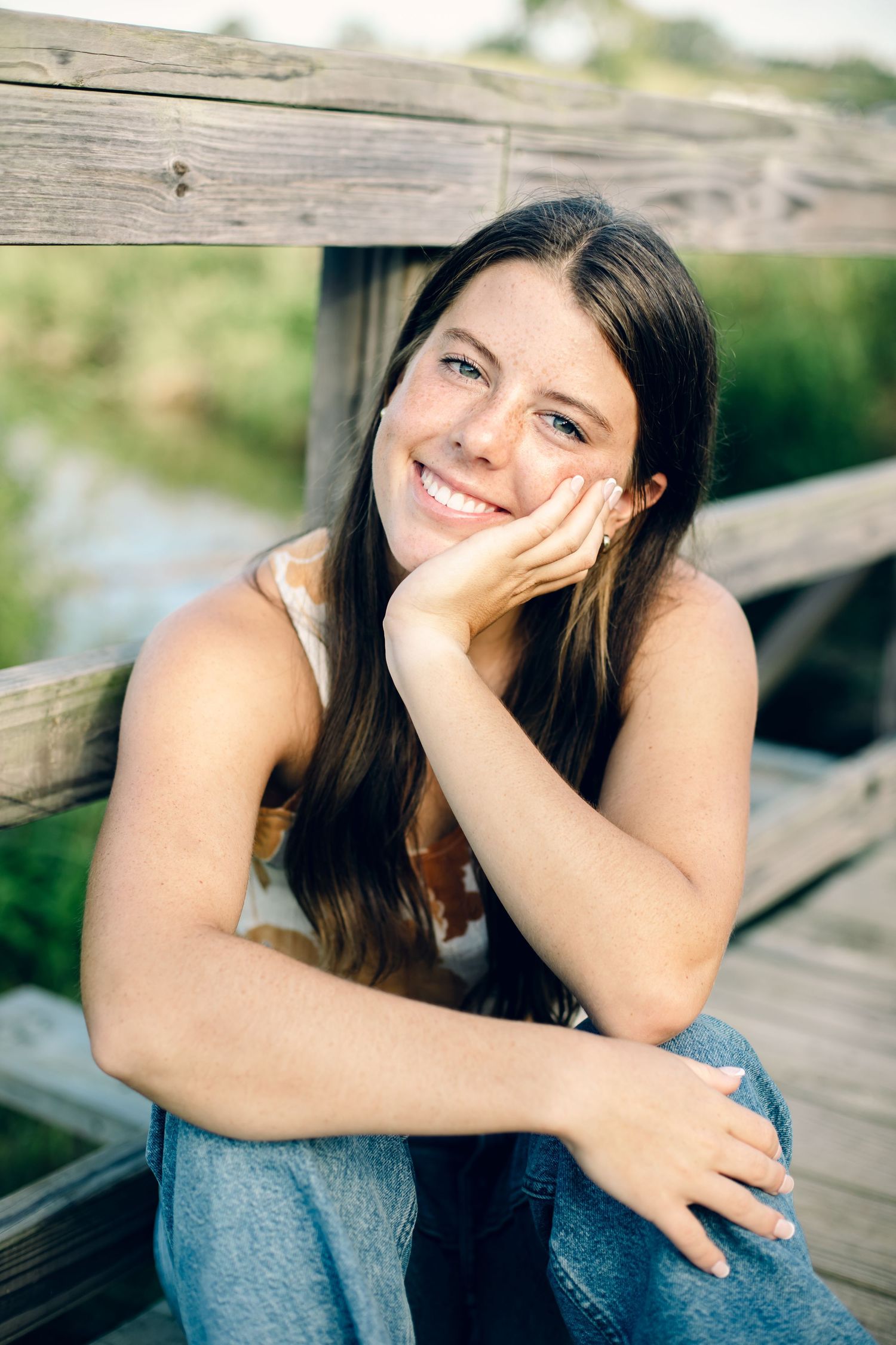 Young woman with long dark hair smiling at camera while sitting casually on wooden structure outdoors in natural setting.