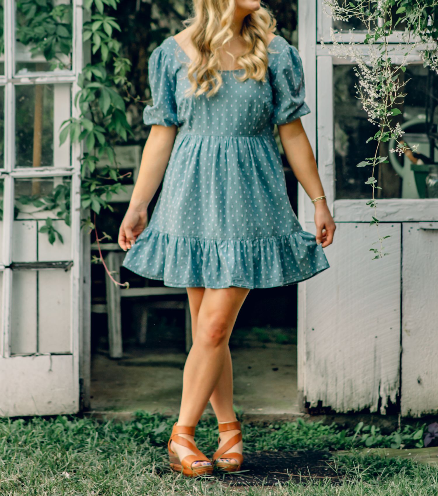 Woman in blue polka dot ruffle mini dress with puff sleeves and tan wedge sandals standing in garden doorway.