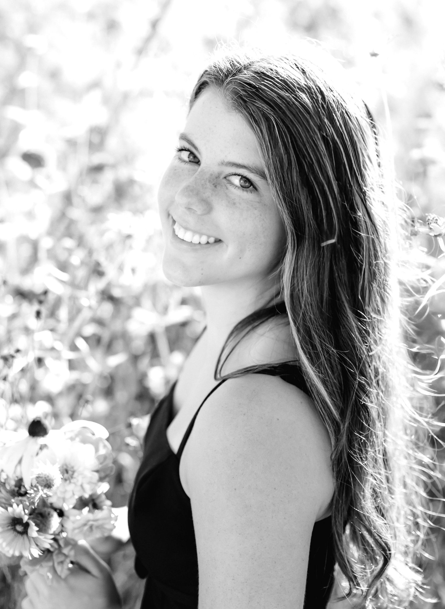 Black and white portrait of a smiling young woman with long hair holding flowers outdoors.