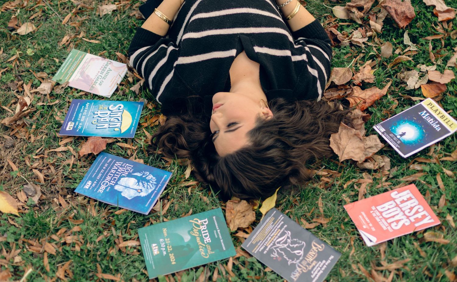 Young woman lying on grass surrounded by colorful books and autumn leaves in an outdoor reading scene.