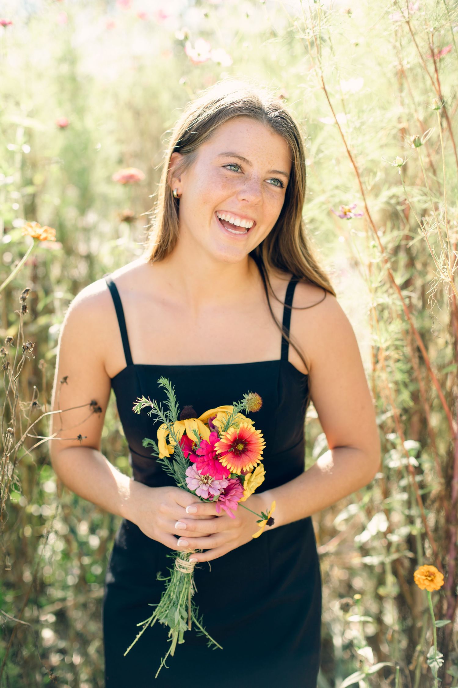 Smiling young woman in black dress holding colorful wildflower bouquet outdoors in sunlit meadow.