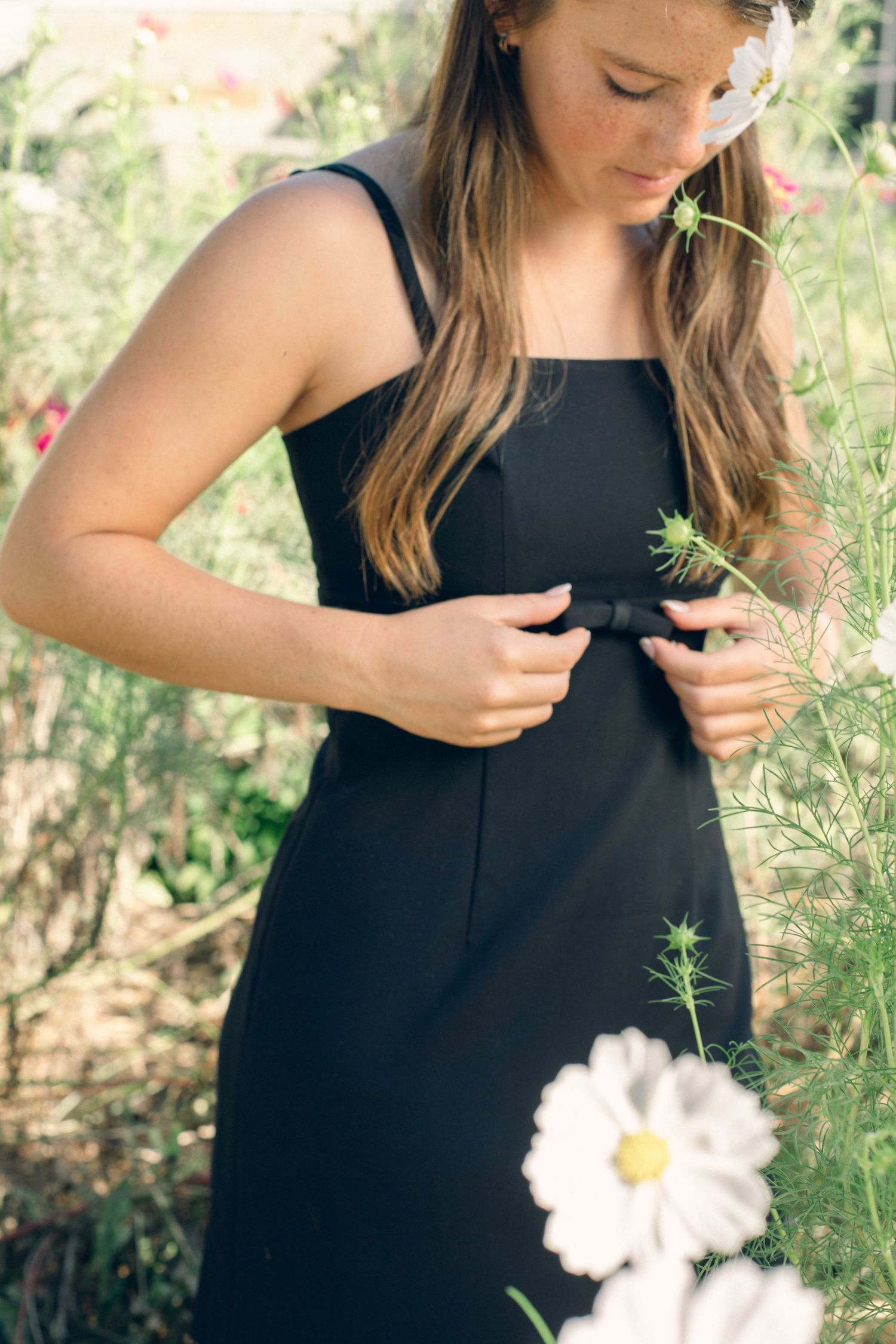 Young woman in black sundress holding a flower stem, surrounded by white cosmos blooms in a garden.