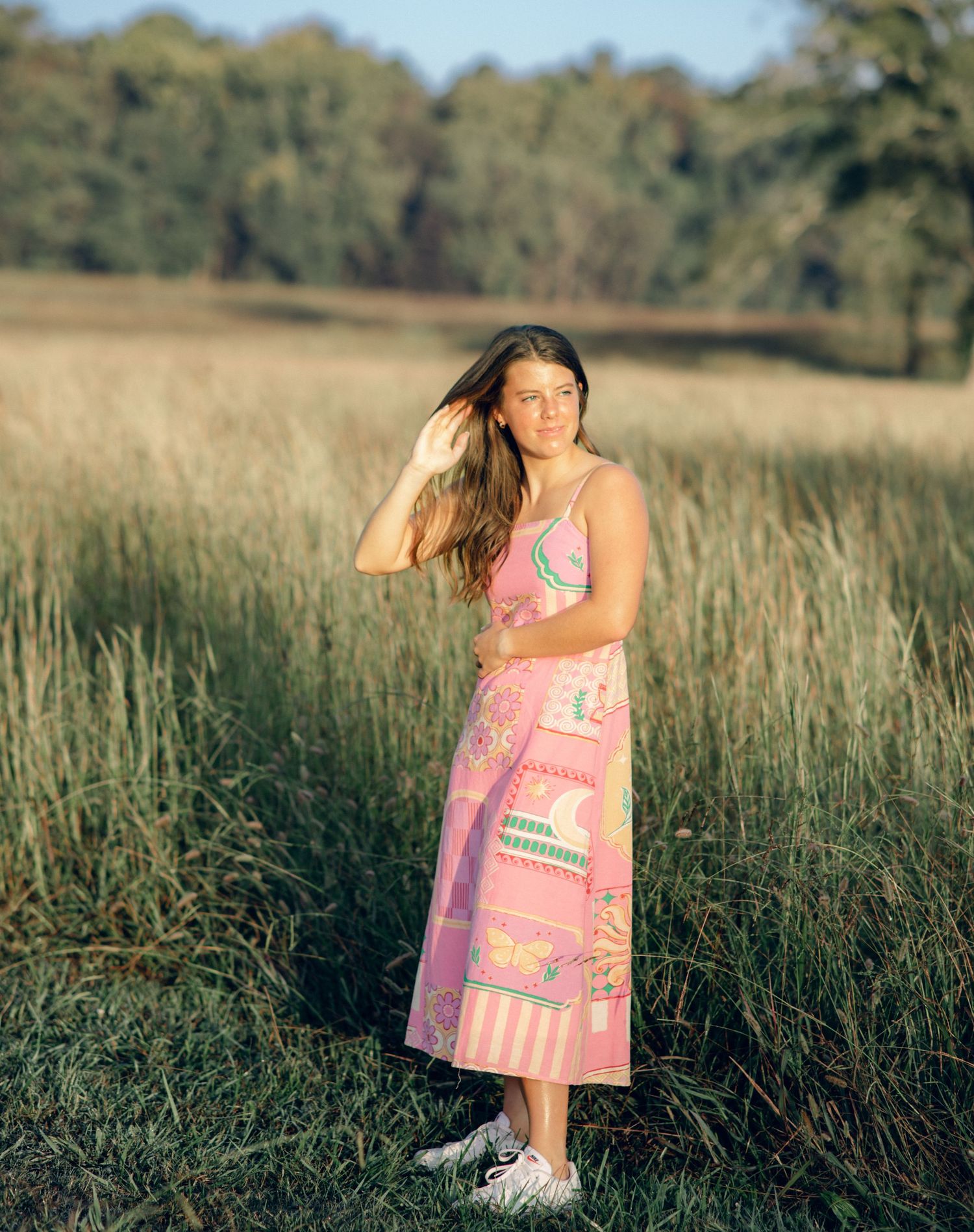 Woman in pink patterned dress standing in grassy field with trees in background during golden hour.