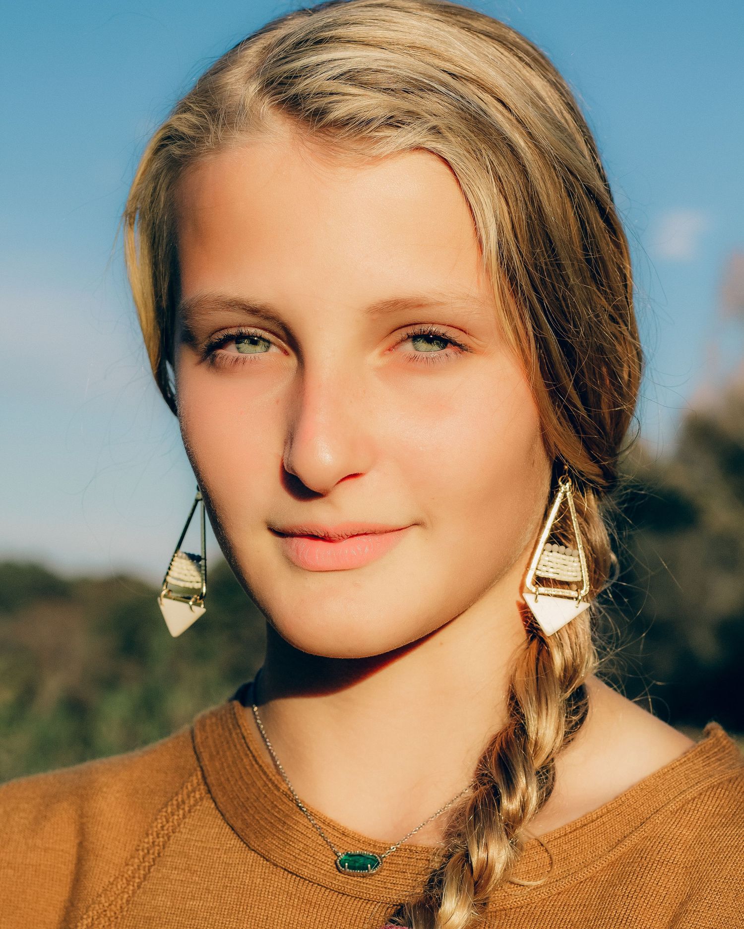 Young woman with blonde braided hair wearing gold geometric earrings and green pendant necklace outdoors.
