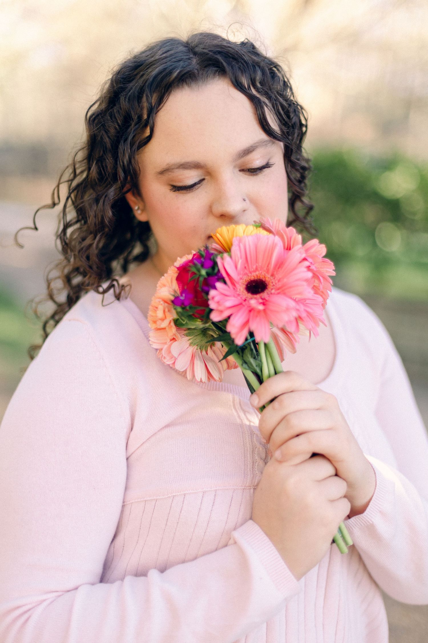 Young woman in pink dress smelling a bouquet of colorful gerbera daisies outdoors in a garden setting.