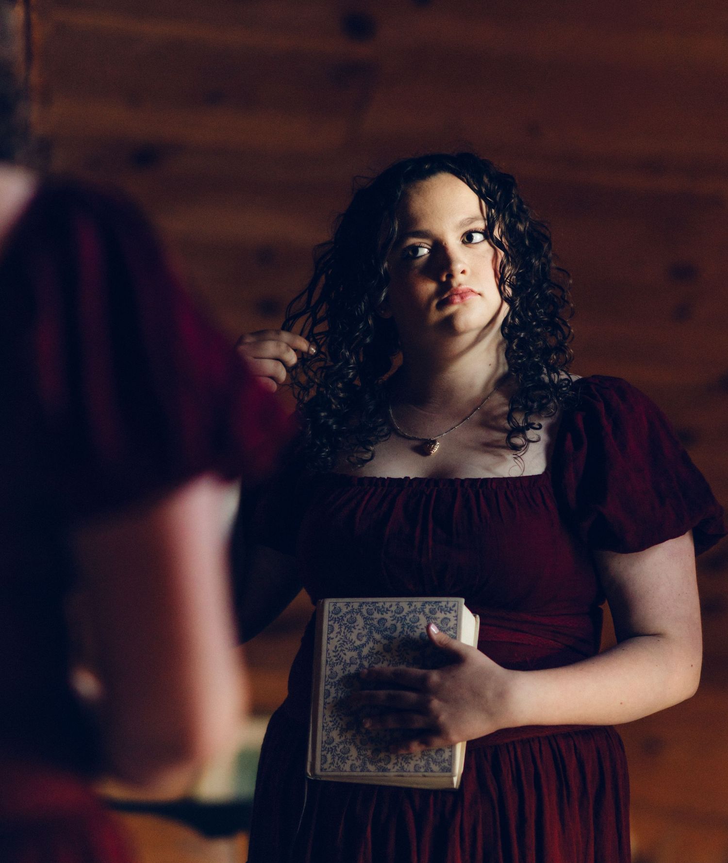 Ashlyn in red velvet dress holding a book, with curly dark hair, standing in a rustic wooden room.