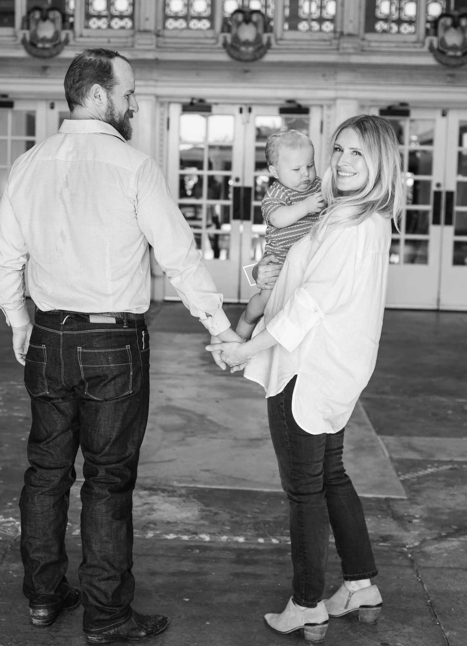 Black and white photo of a couple holding hands with a baby at an elegant indoor venue.