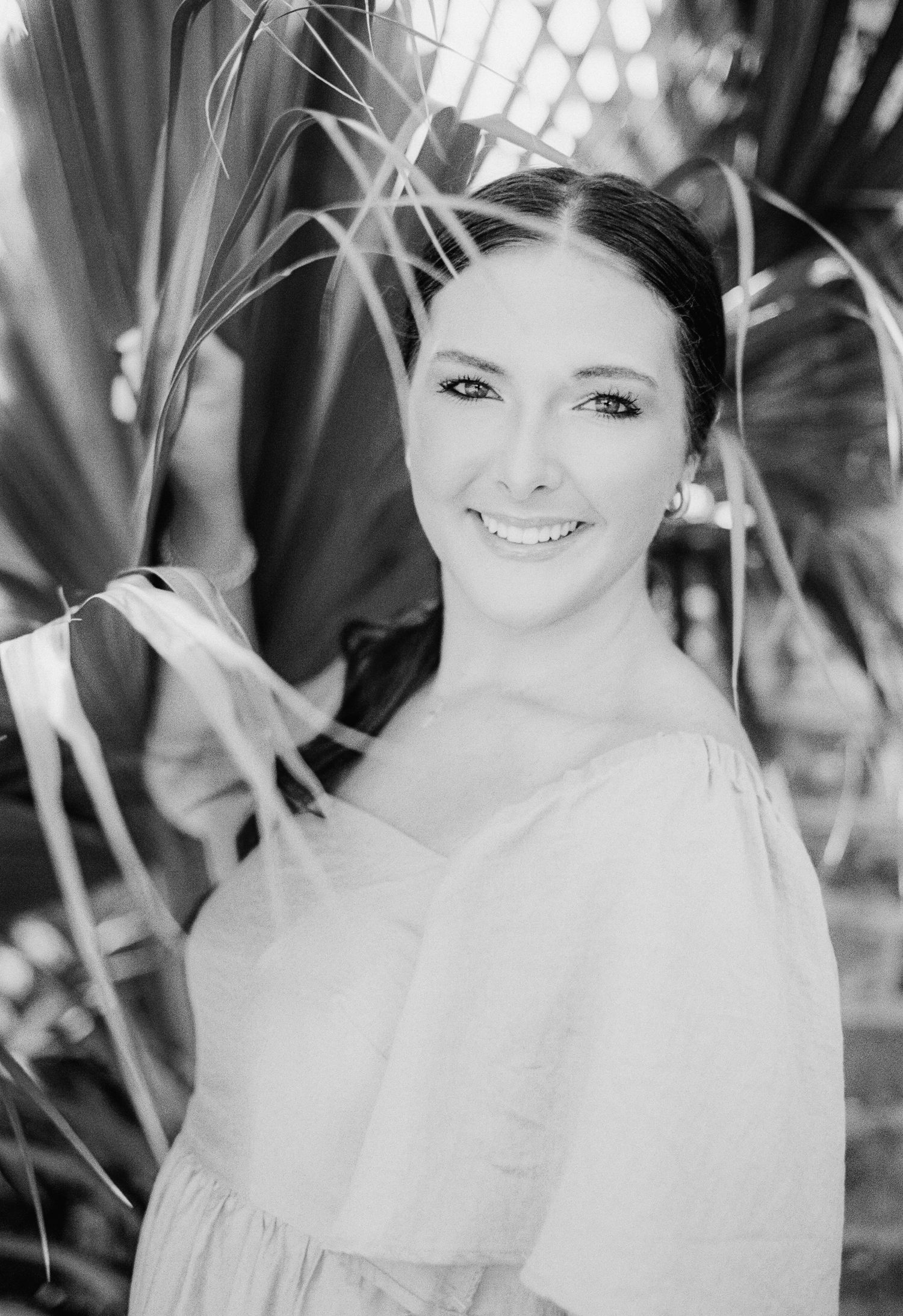Black and white portrait of a smiling young model in a  off-shoulder top surrounded by tropical palm leaves in downtown Charleston, SC.