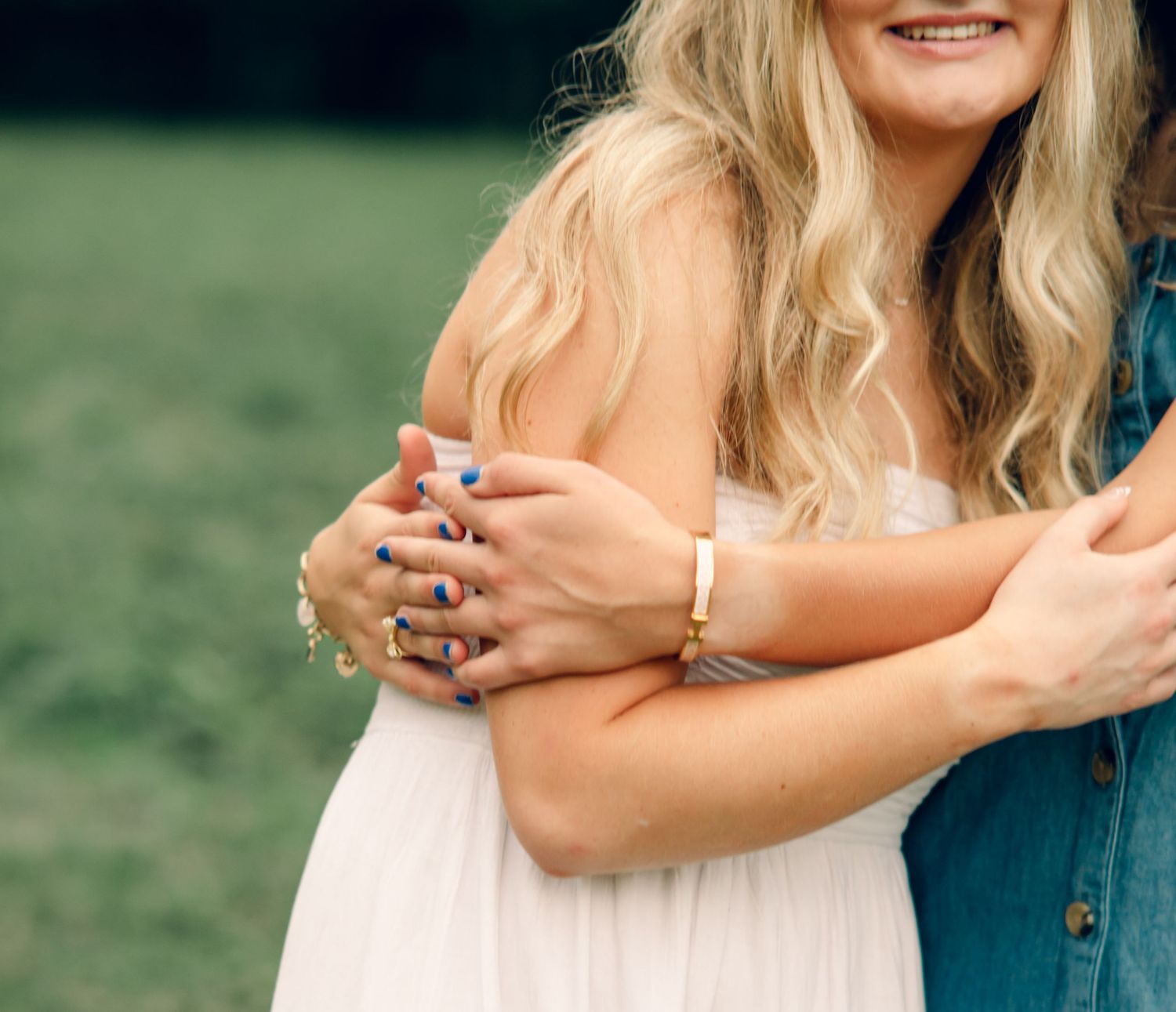 Two people share a warm embrace outdoors, with the woman wearing rings and a gold bracelet, dressed in a white dress.