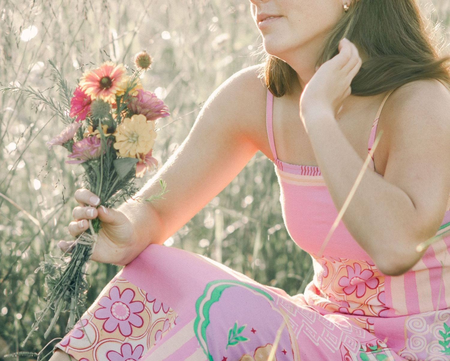 Young woman in pink floral dress holding wildflowers while sitting outdoors in a field.
