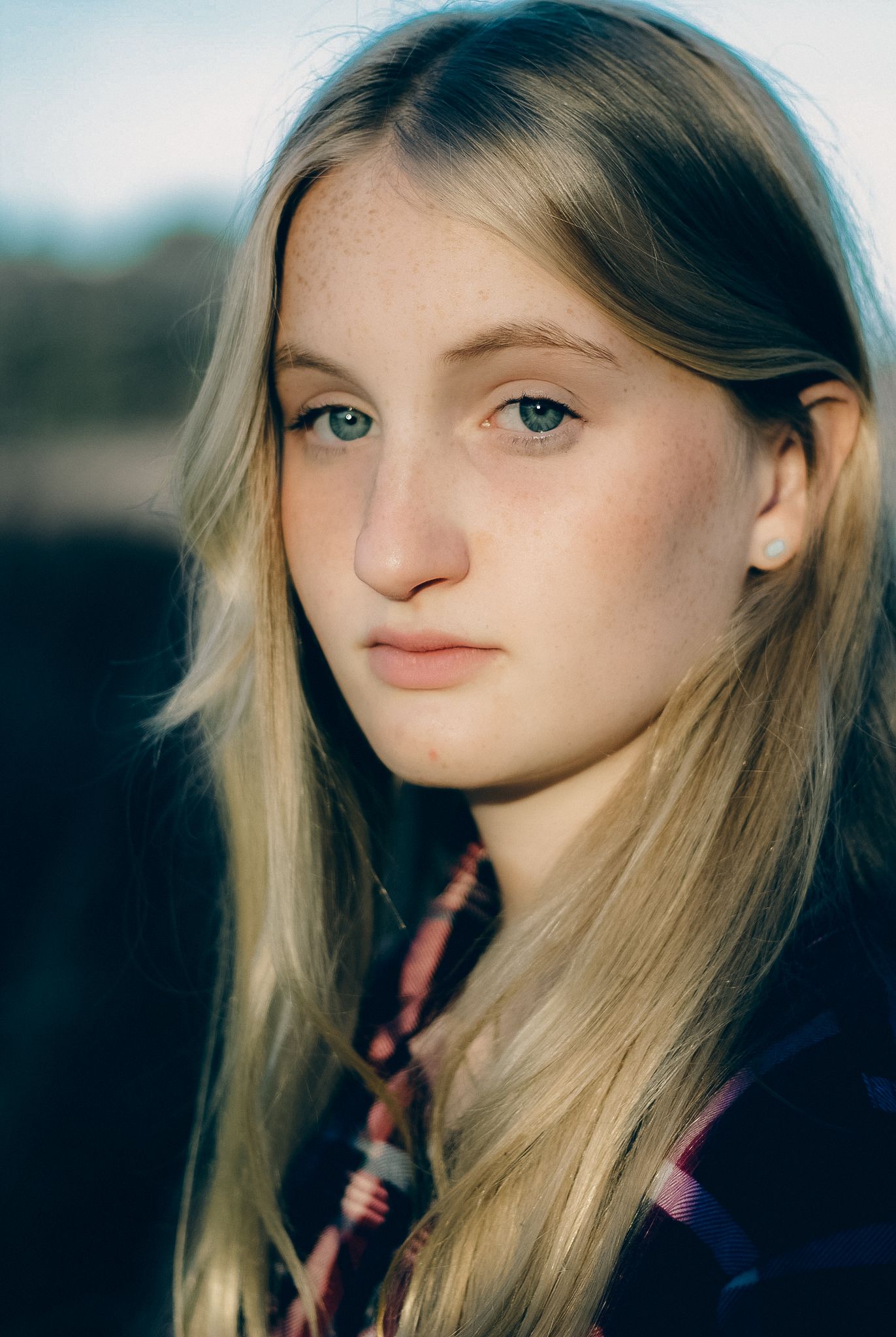 Close-up portrait of a young blonde woman with blue-green eyes and freckles wearing a plaid shirt outdoors.